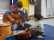Therapeutin mit Mund-Nasen-Schutz spielt Gitarre, Junge sitzt vor ihr und zupft an einer Gitarre, die auf dem Boden liegt.
