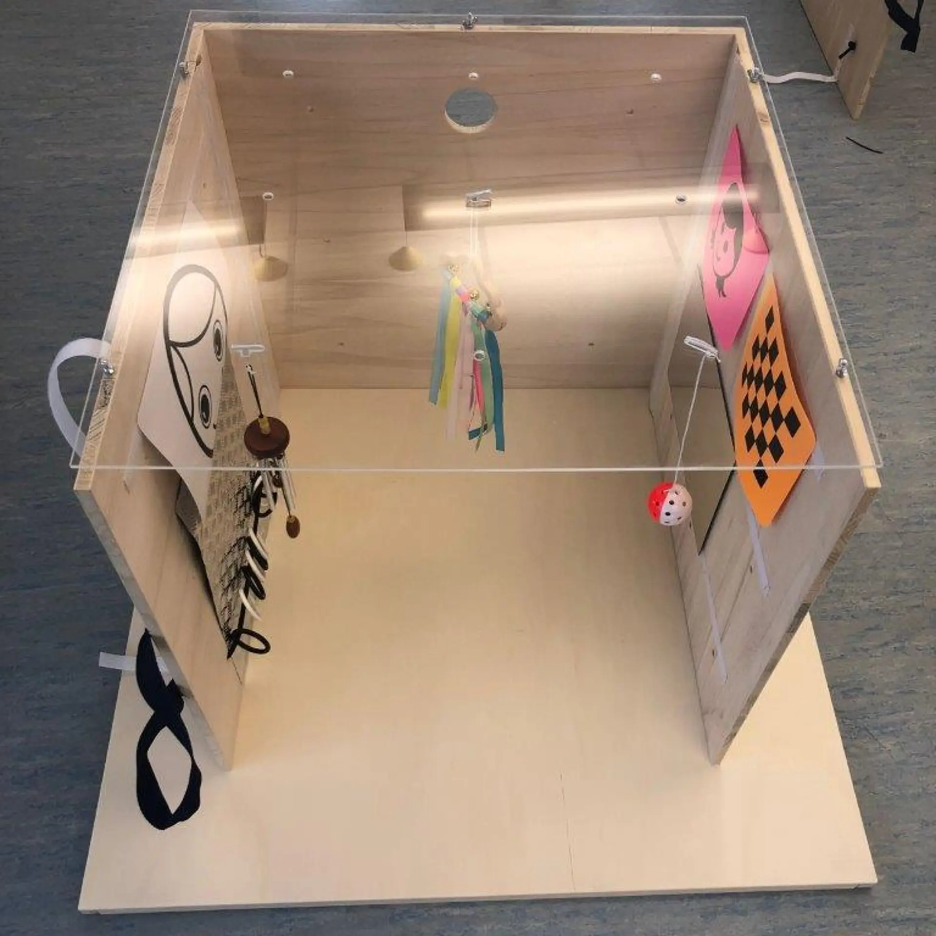 Ein pädagogisches Entwicklungsspielgerät, das einer Holzkiste mit transparentem Deckel ähnelt. Innen hängen verschiedene taktile Materialien, darunter bunte Bänder, eine Kette mit Schlüsselring und kontrastreiche visuelle Karten (schwarz-weißes Gesicht, orange-schwarz karierte Flagge). Das Gerät dient zur Exploration und Stimulation in der Frühförderung.