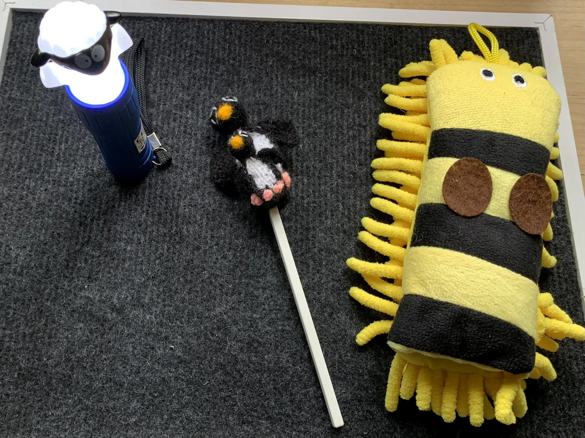 Drei taktile Spielzeuge für die Frühförderung auf einer dunklen Oberfläche: Eine leuchtende Taschenlampe mit einem Schafskopf, eine Rassel mit zwei kleinen, gestrickten Pinguinen und ein Fühl- und Greifspielzeug mit Fransen.