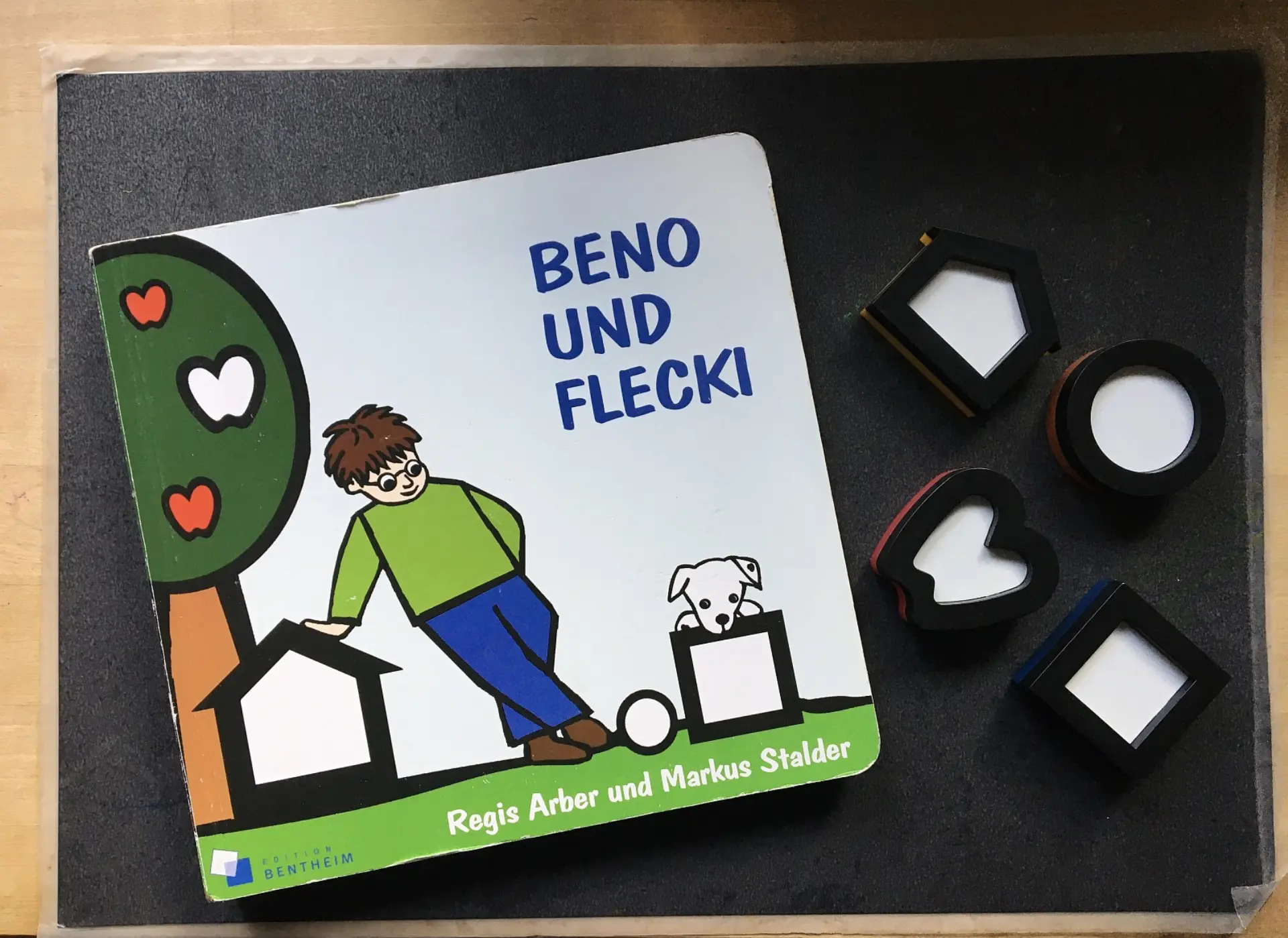 Kinderbuch "Beno und Flecki" von Regis Arber und Markus Stalder, liegend neben vier kontrastreichen Spielsteinen in geometrischen Formen (Quadrat, Kreis, Pentagon und Herz). Das Buchcover zeigt einen Jungen, einen Baum mit Herzen und einen Hund.