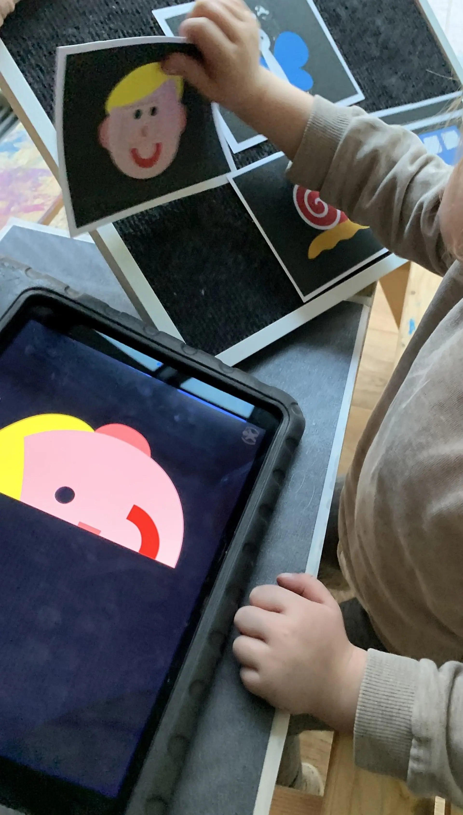Nahaufnahme aus der Frühförderung: Ein Kind (nur Hände und Schulter sichtbar) hält eine laminierte Karte mit einem einfachen, bunten Gesichtsmotiv. Die Karte wird über ein Tablet gehalten, dessen Bildschirm eine stark vergrößerte, kontrastreiche und farbreduzierte Darstellung desselben Motivs anzeigt. Das Material dient zur Förderung der visuellen Wahrnehmung und des Erkennens.