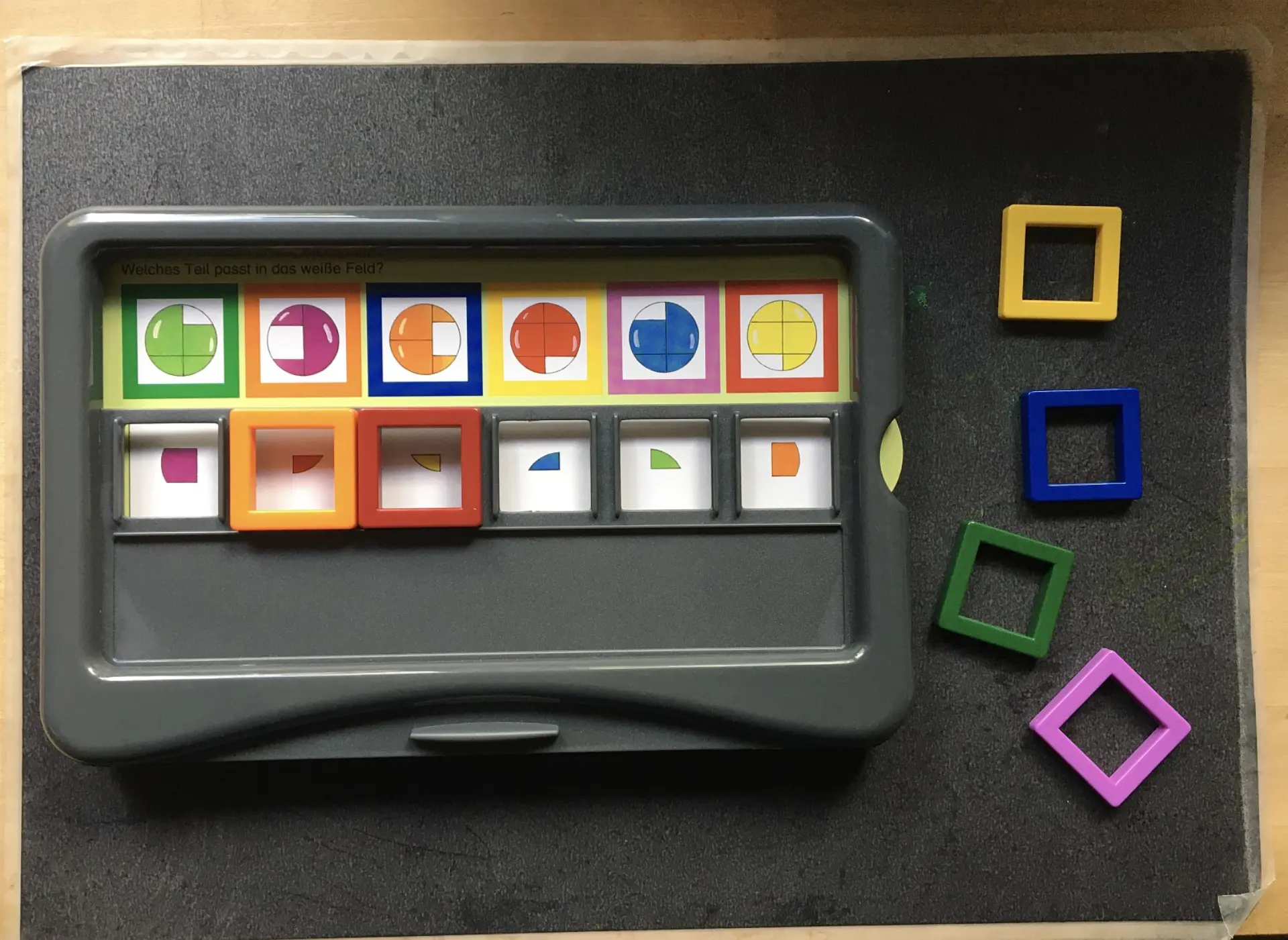 Lernspiel zur visuellen Wahrnehmung und Formerkennung, liegend auf einer dunklen Oberfläche. Das Spielbrett zeigt bunte Bilder mit geometrischen Formen und hat eine Reihe von Fächern, in denen farbige Rahmen und kleine Formteile liegen. Rechts liegen vier quadratische, farbige Rahmen.