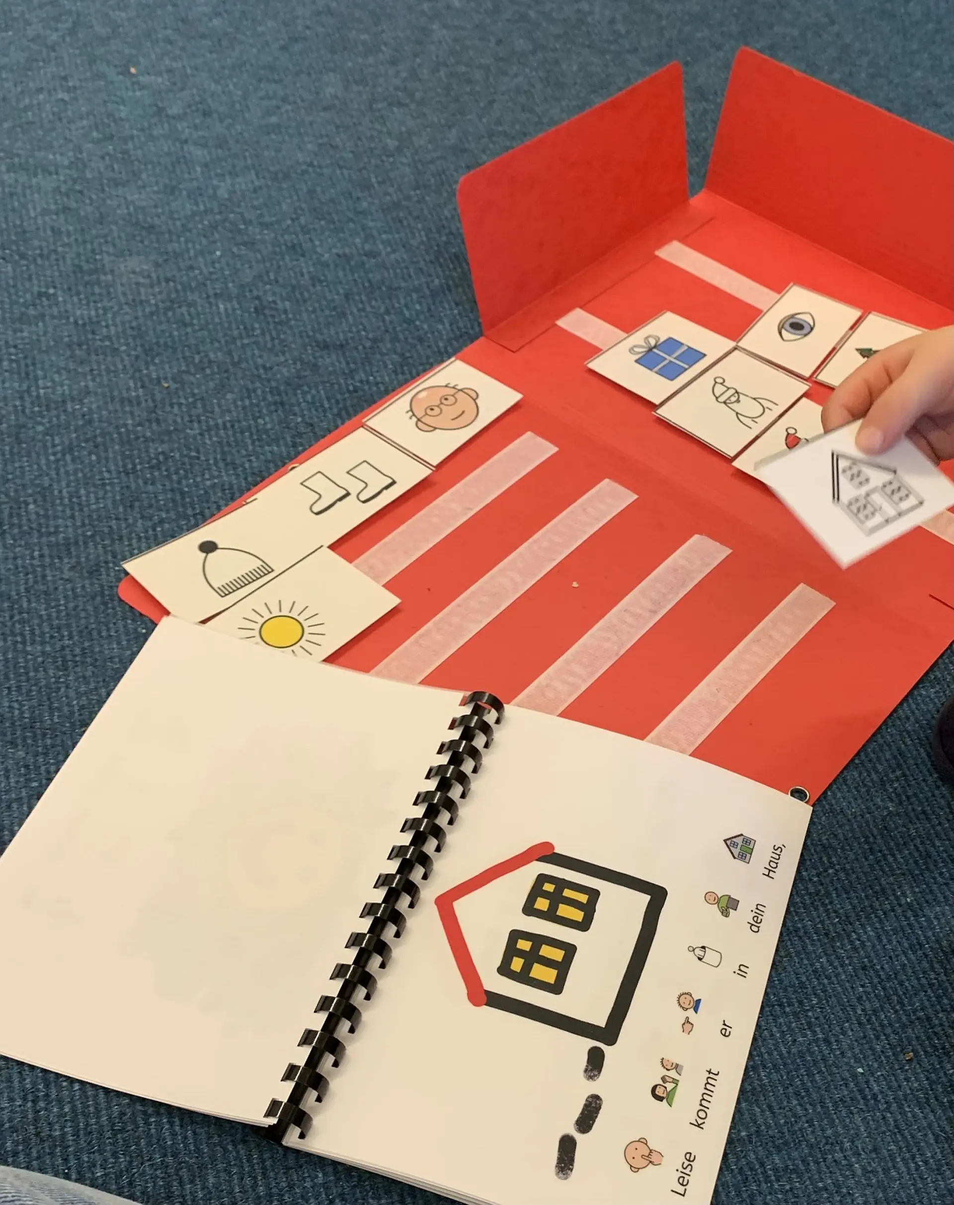 Eine geöffnete Papiermappe liegt auf dem Boden. Dazu gibt es noch aufgeblättertes Arbeitsheft mit einer einfachen Zeichnung von einem Haus. Eine kleine Hand hält ein Kärtchen mit dem selben Haus darauf und soll es an die richtige Stelle in der Mappe hinlegen. In der Mappe liegen schon weitere Karten, die zu diesem Lernset der Frühförderung gehören.