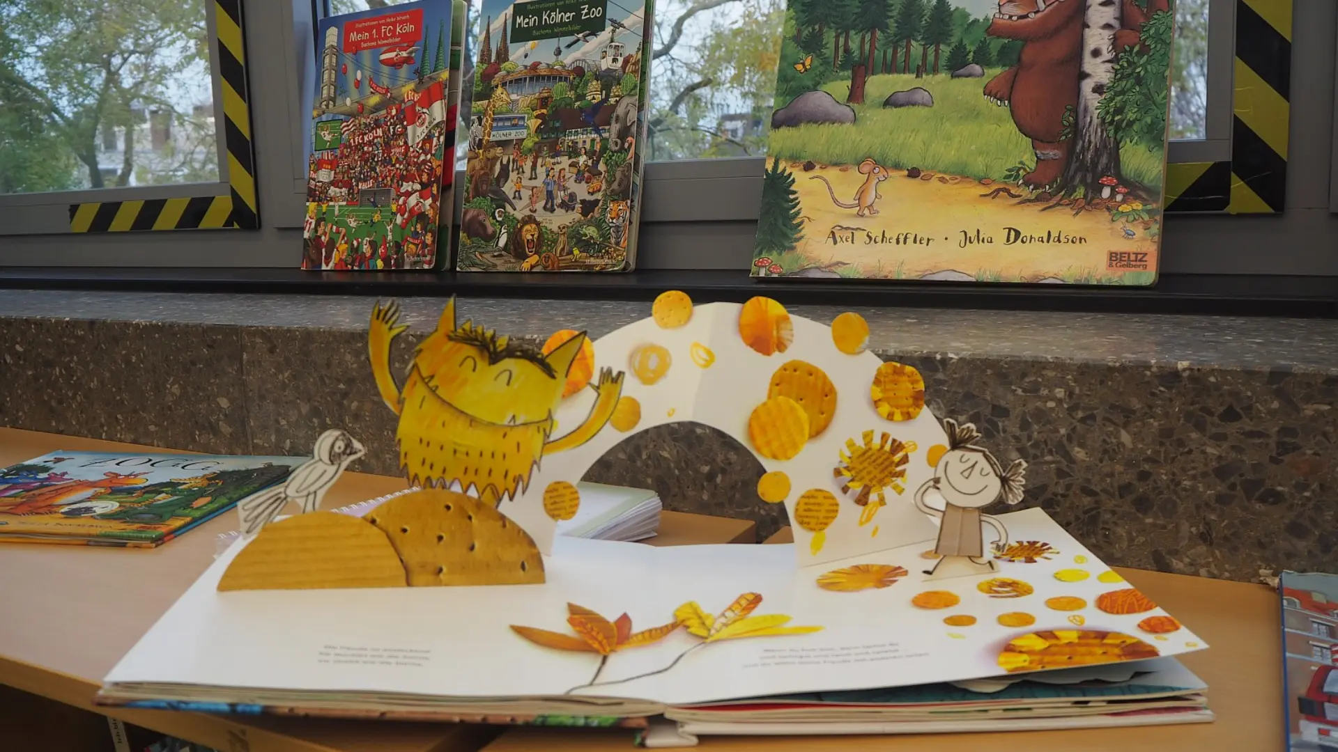 Auf einem Fensterbrett stehen drei große Bilderbücher, darunter "Der Grüffelo" von Axel Scheffler und Julia Donaldson. Im Vordergrund liegt ein weiteres Pop-up-Bilderbuch mit einer gelben Monsterfigur und herbstlichen Motiven geöffnet auf einem Holztisch.
