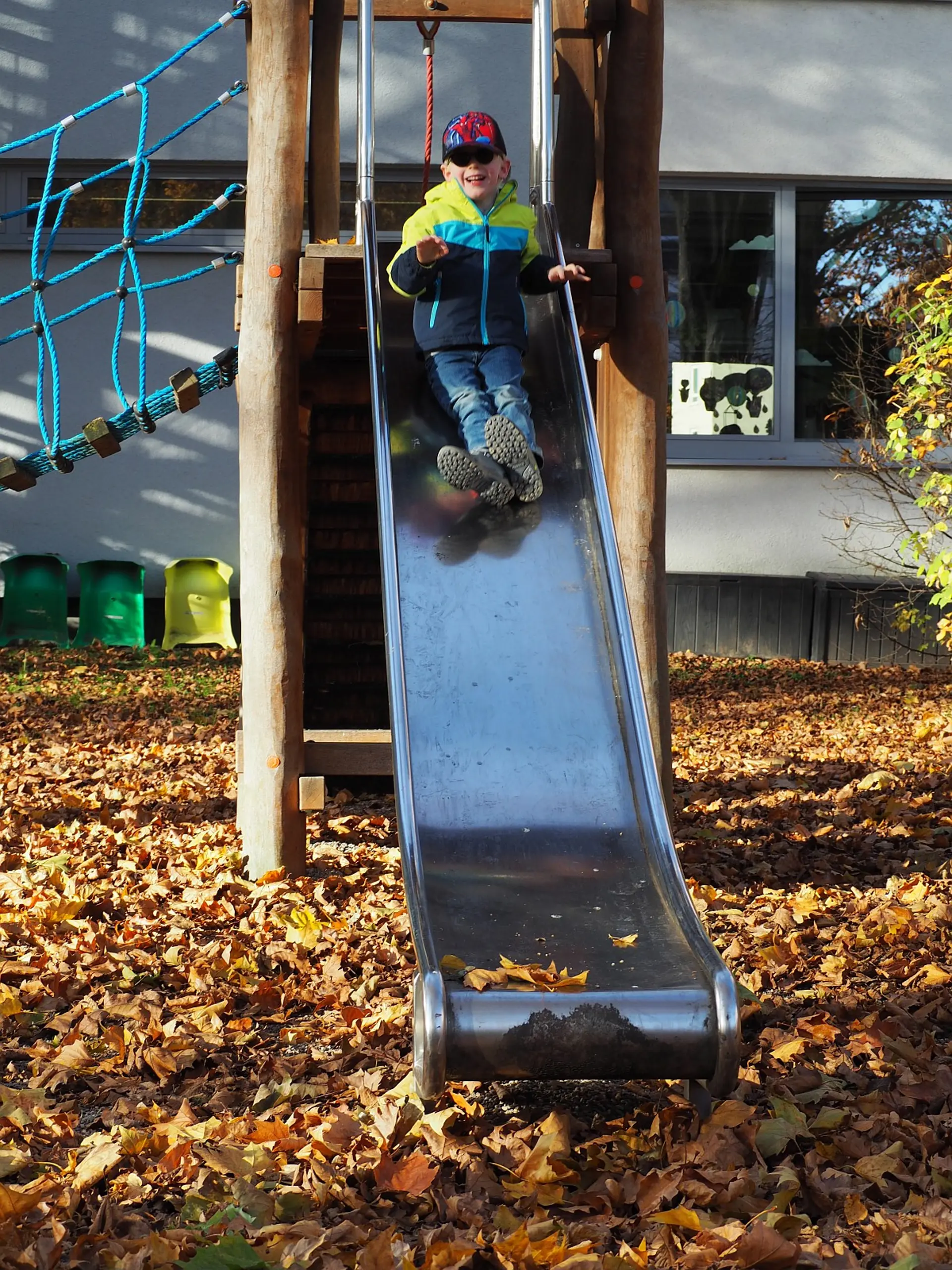 Ein Kind im Vorschulalter mit dunkler Brille rutscht an der Rusche im Schulinnenhof herunter. Dabei lacht es offensichtlich vergnügt.