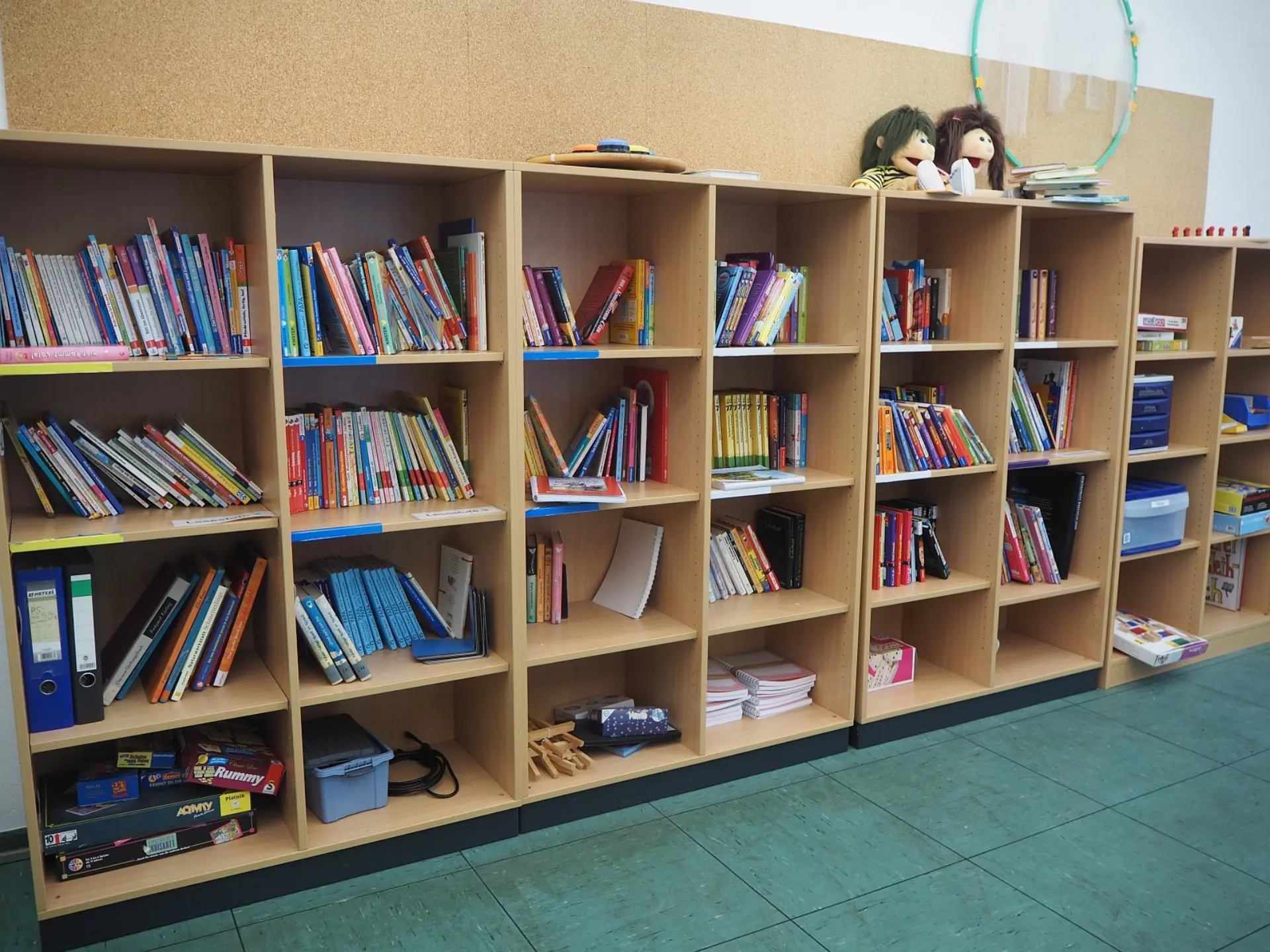 Mehrere Reihen von hellbraunen Holzregalen, gefüllt mit Kinderbüchern mit bunten Buchrücken. Die Regale stehen in einer Schulbücherei auf einem türkisgrünen Boden. Oben auf den Regalen stehen Brettspiele, einige Bücherstapel und zwei Plüschpuppen mit langen, dunklen Haaren.