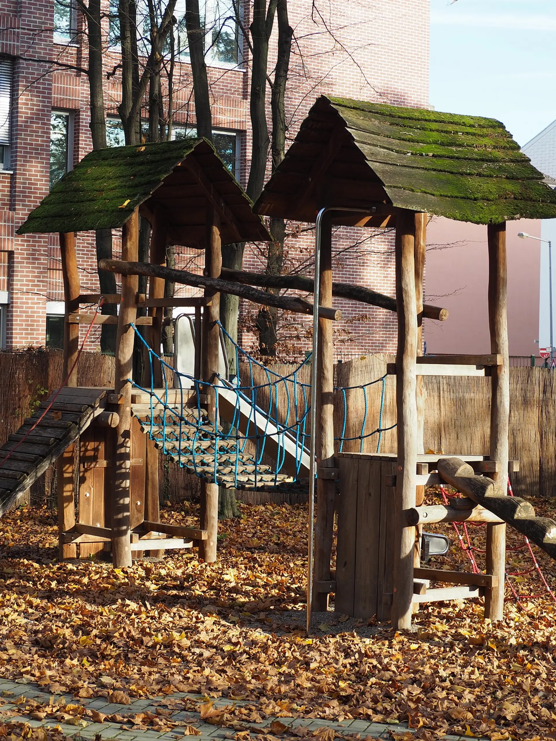 Ein stabiles Spielgerät aus Holz auf dem Schulhof. Es besteht aus zwei Türmen mit bemoosten Dächern, die durch eine Hängebrücke mit Seilnetz verbunden sind. Der Boden um das Gerüst ist mit trockenem Herbstlaub bedeckt. Im Hintergrund ist eine Backsteinfassade sichtbar.