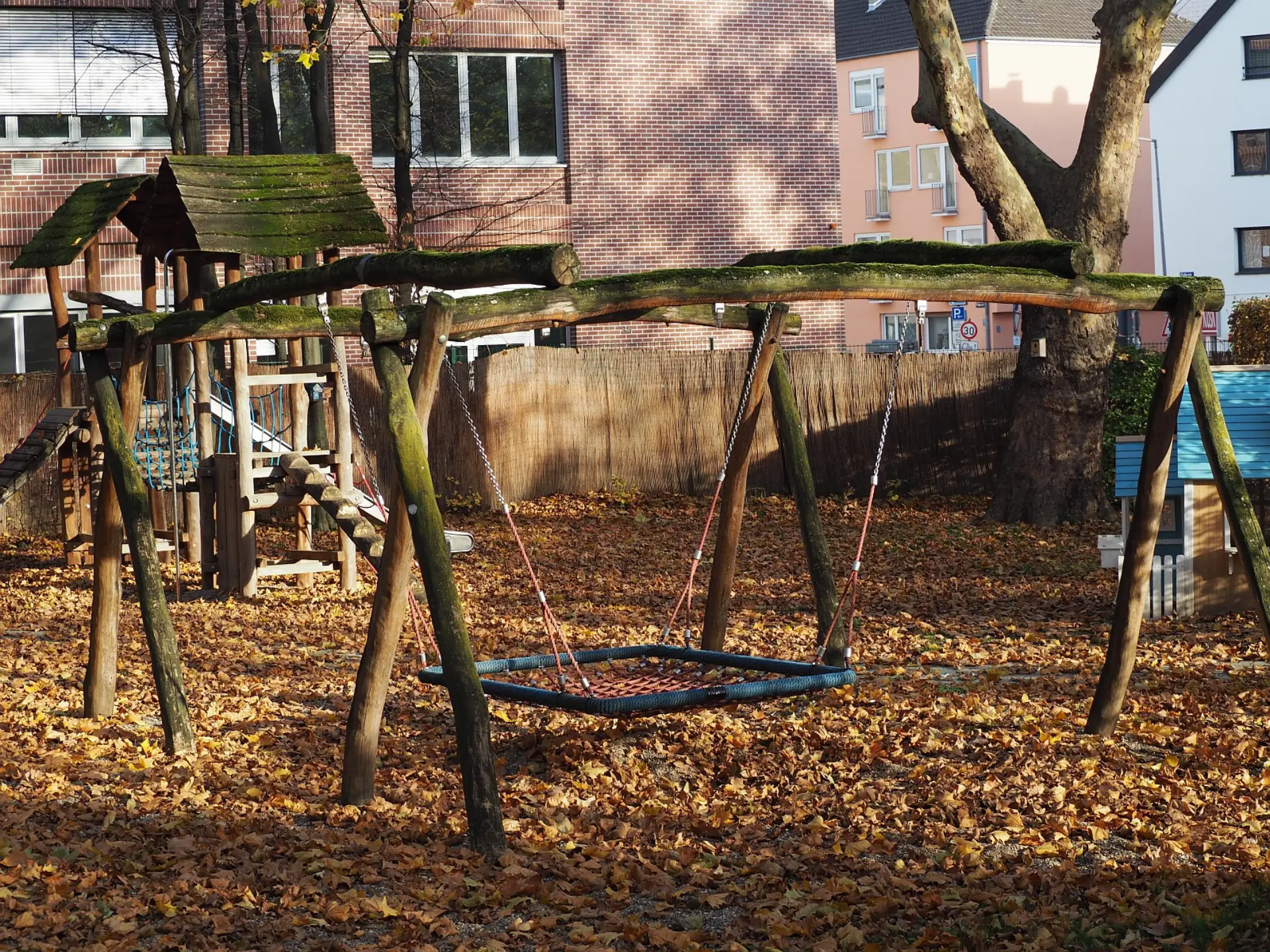 Ein Spielbereich auf dem Schulhof im Herbst. Das Bild zeigt eine große Schaukelanlage aus rustikalem, bemoostem Holz mit einer breiten, quadratischen Netzschaukel. Der Boden ist vollständig mit trockenem Laub bedeckt. Im Hintergrund sind ein Holzklettergerüst und mehrstöckige Backsteingebäude sichtbar.