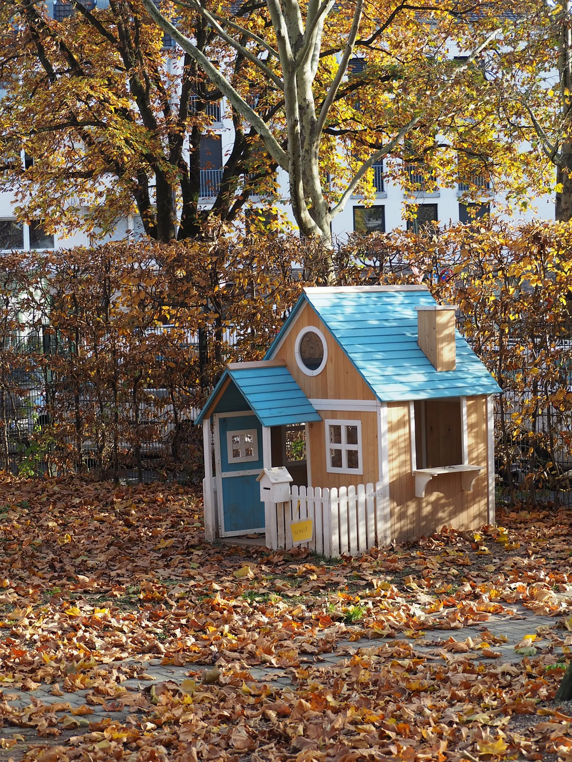 Ein Holz-Spielhaus mit hellblauem Dach, einer kleinen Veranda und einem Lattenzaun auf dem Schulhof. Es steht auf einer Fläche, die mit Herbstlaub bedeckt ist. Im Hintergrund säumen eine hohe Hecke und Bäume den Spielbereich.