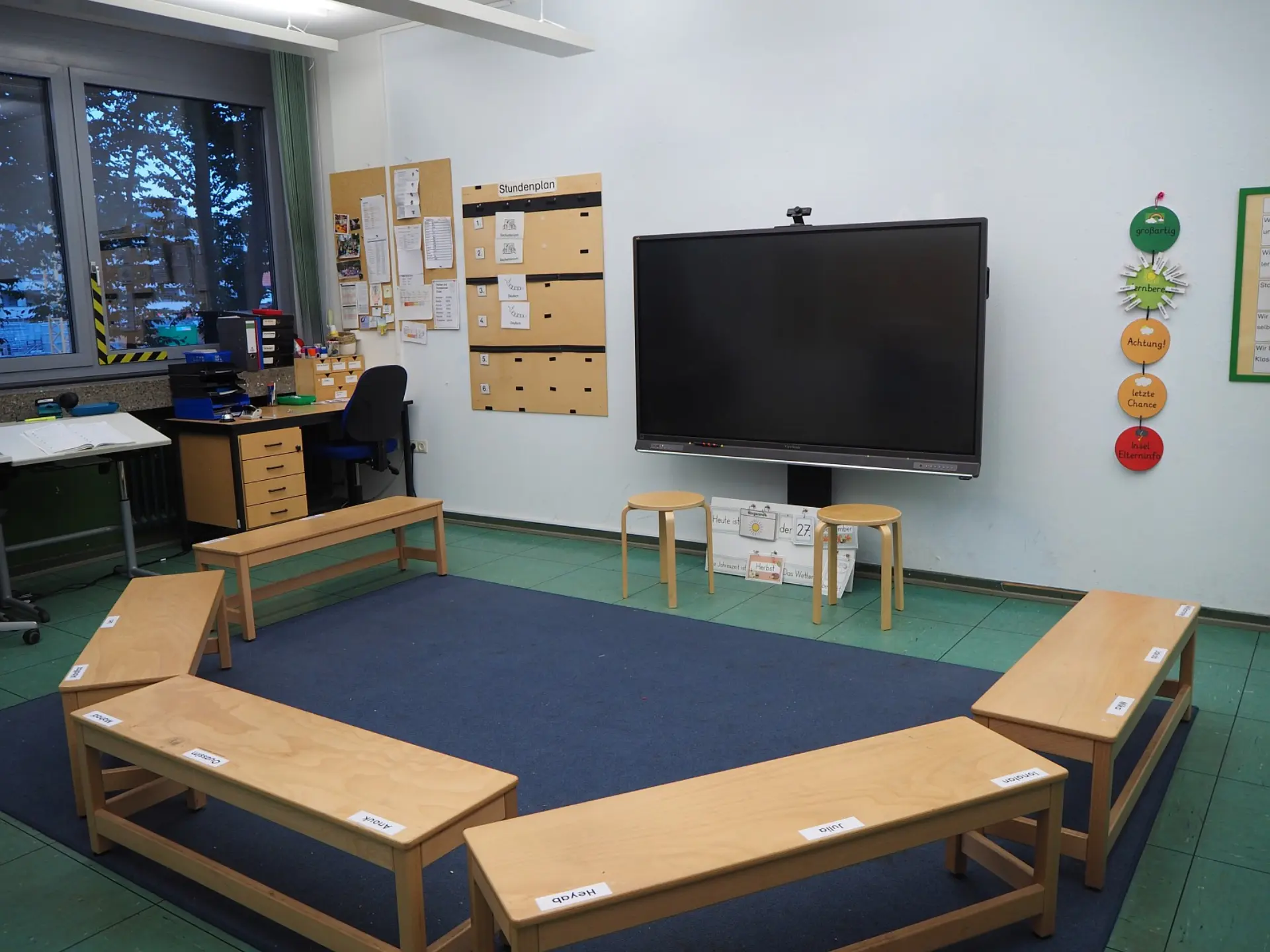 Ein Klassenraum der LVR-Severin-Schule, eingerichtet für inklusiven Unterricht. In der Mitte des Raumes stehen Holzbänke in U-Form auf einem Teppich vor einem interaktiven Bildschirm. Links befindet sich ein Schreibtisch, rechts hängen Werte-Symbole an der Wand.