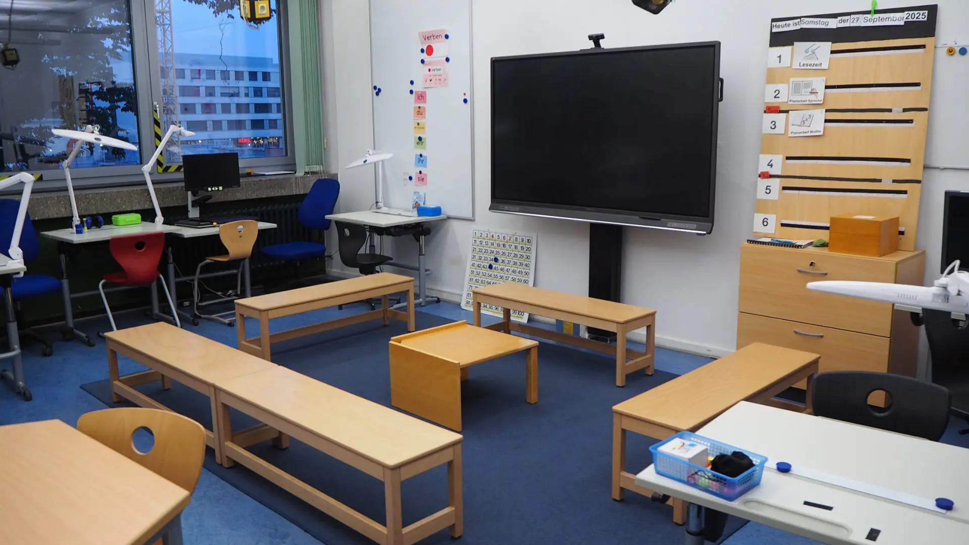 Ein moderner Klassenraum der LVR-Severin-Schule. Er verfügt über ein interaktives Display, eine flexible Sitzgruppe aus Holzbänken auf einem Teppich, Einzelschreibtische am Fenster mit Lampe für Sehbeeinträchtigte sowie einen gut organisierten Schrank mit Lesematerialien.