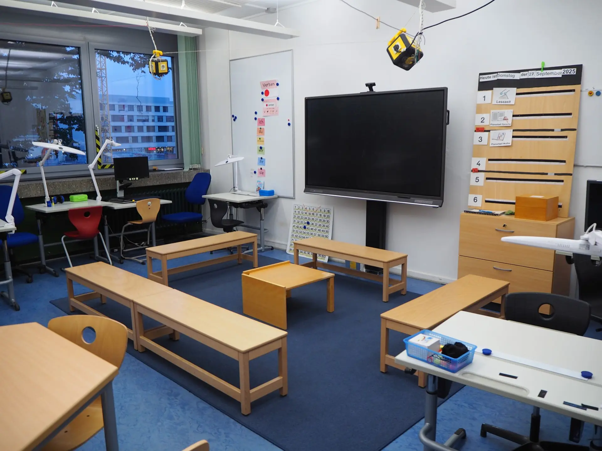 Ein moderner Klassenraum der LVR-Severin-Schule. Er verfügt über ein interaktives Display, eine flexible Sitzgruppe aus Holzbänken auf einem Teppich, Einzelschreibtische am Fenster mit Lampe für Sehbeeinträchtigte sowie einen gut organisierten Schrank mit Lesematerialien.