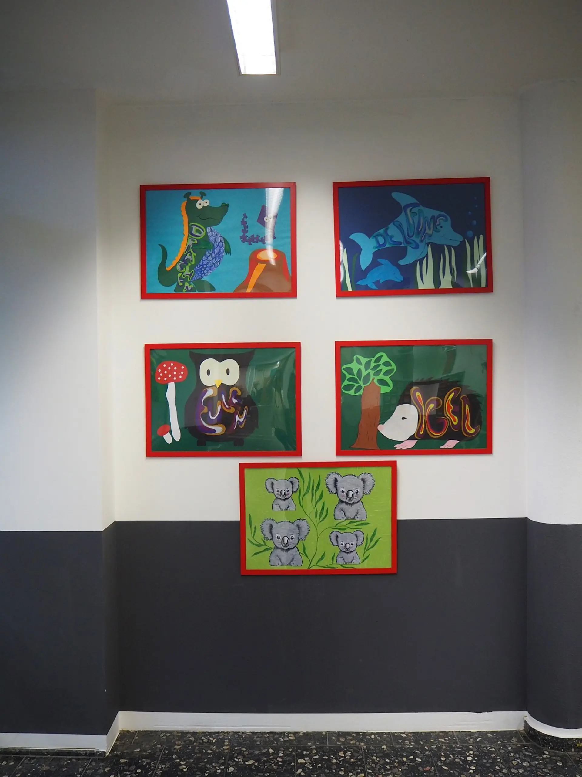 Wandgestaltung im Schulflur mit fünf bunten, gerahmten Illustrationen von Klassentieren: Oben links ein Krokodil, rechts Delfine; darunter eine Eule und ein Igel; mittig unten vier Koalas. Die Bilder hängen im Schulflur.