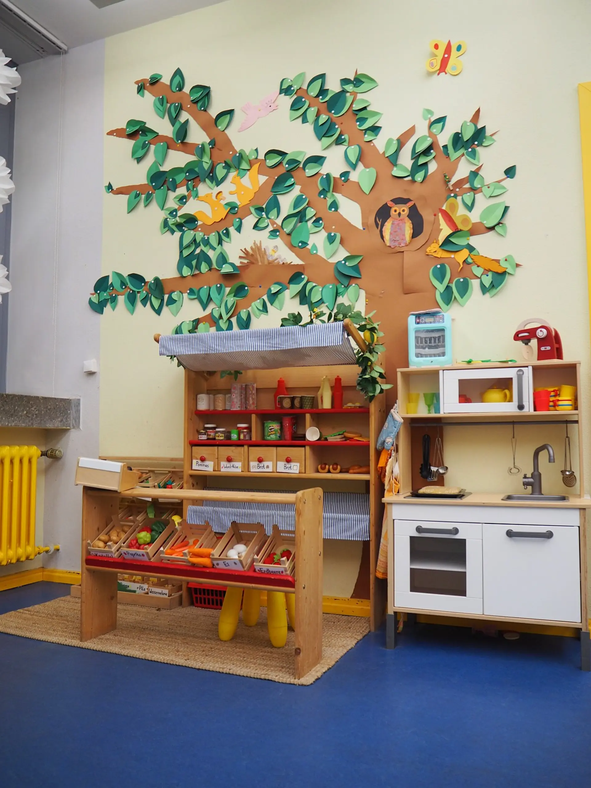 Ein fantasievoll gestalteter Spielbereich in der Offenen Ganztagsschule (OGS). Das Set besteht aus einem hölzernen Marktstand mit Obst und Gemüse sowie einer Spielküche mit Spüle und Mikrowelle. Die Wand darüber ist mit einer großen, bunten Bastelarbeit in Form eines Baumes mit Blättern und Tieren (Eule, Eichhörnchen) dekoriert.