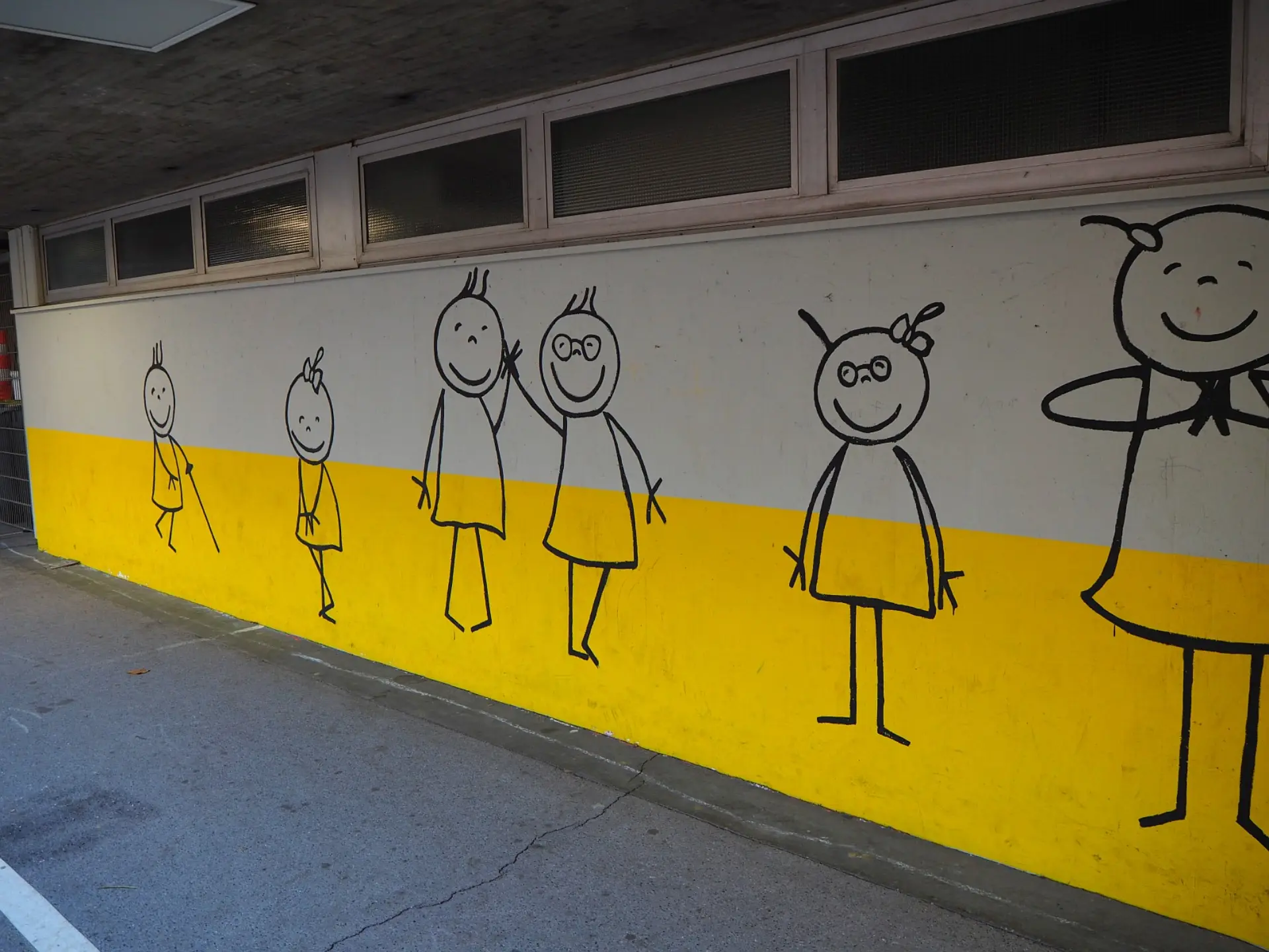Detaillierte Wandillustrationen auf dem Schulgelände der LVR-Severin-Schule. Die Zeichnung zeigt fröhliche, gezeichnete Kinderfiguren in schwarzer Farbe auf einem weiß-gelb geteilten Hintergrund unter einem Betondach.