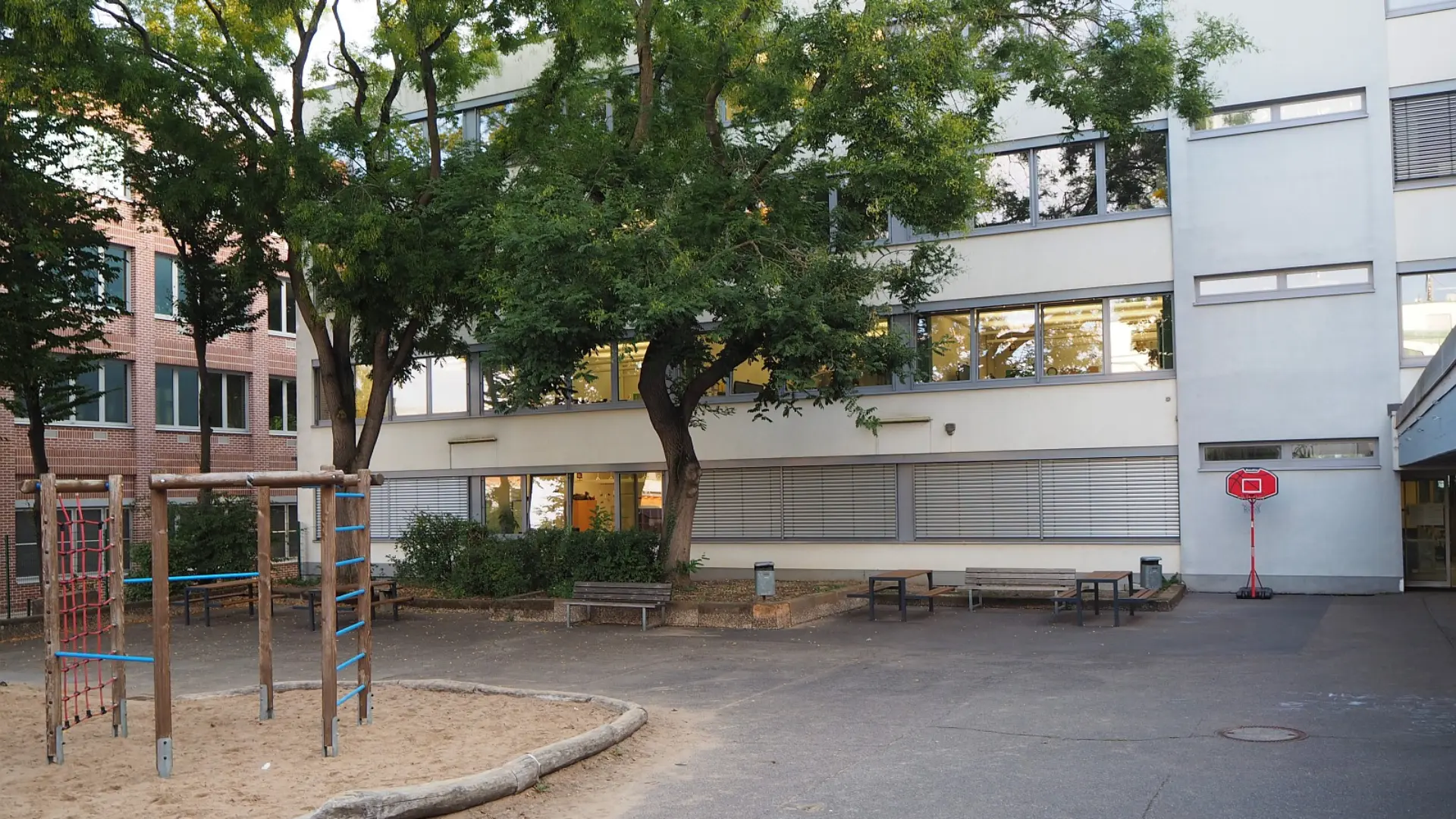 Blick auf das Hauptgebäude der LVR-Severin-Schule. Im Vordergrund ist ein Teil des Schulhofs mit einem Sandkasten, einem Klettergerüst und Bänken unter Bäumen zu sehen. Rechts befindet sich ein Basketballkorb.