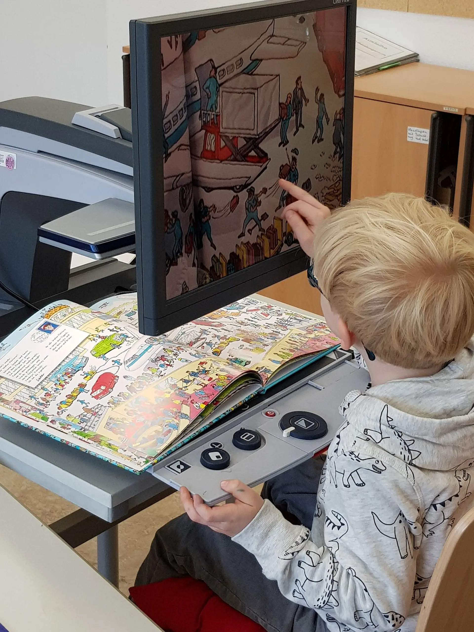 Ein Kind im Grundschulalter sitzt am Schreibtisch. Vor ihm ist Bildschirmlesegerät, damit wird ein Buch mit vielen Bildern am Monitor groß angezeigt. Das Kind schaut zum Monitor und drückt ein Element aus dem Bild auf dem Touch-Display.