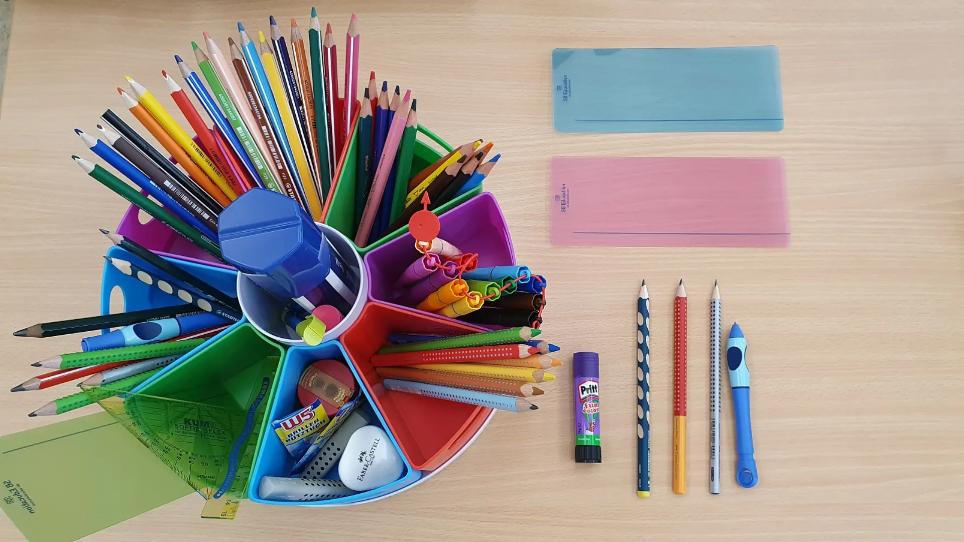 Ein Schreibtisch mit zahlreichen Schreib- und Zeichenutensilien: Im Zentrum ein bunter, runder Stifteorganizer mit einer Fülle von Buntstiften, darunter auch dicke, ergonomische Stifte. Daneben liegen eine Schablone, ein Klebestift, zwei weitere transparenten Schablonen sowie spezielle Dreikant- und ergonomische Bleistifte.