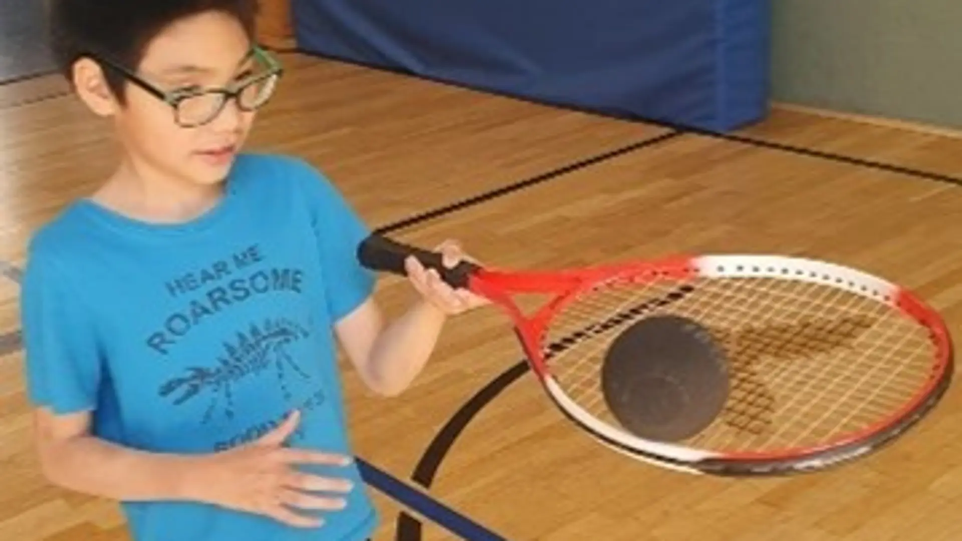 Ein Schüler mit Brille balanciert auf einem Tennisschläger einen Ball für Blinden-Tennis. Er ist in einer Sporthalle.