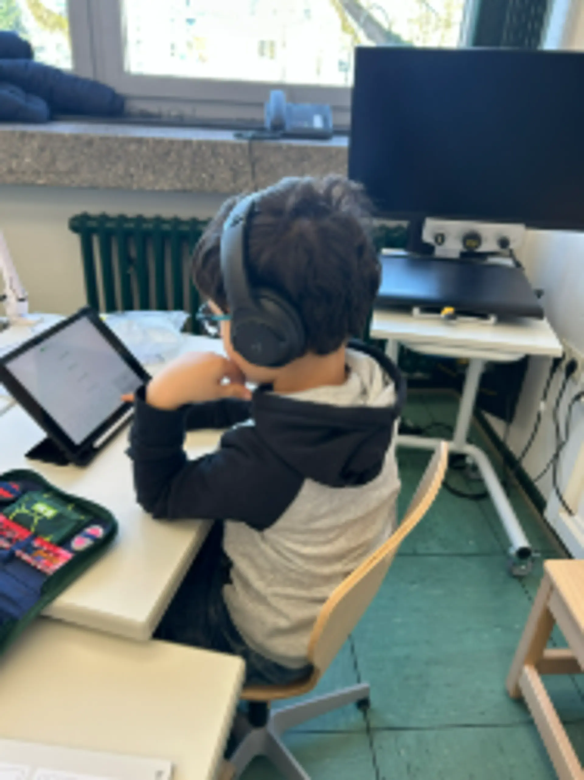 Ein Kind sitzt am Schreibtisch in einem Klassenzimmer, arbeitet am Tablet und hört etwas mit großen Kopfhörern dazu.