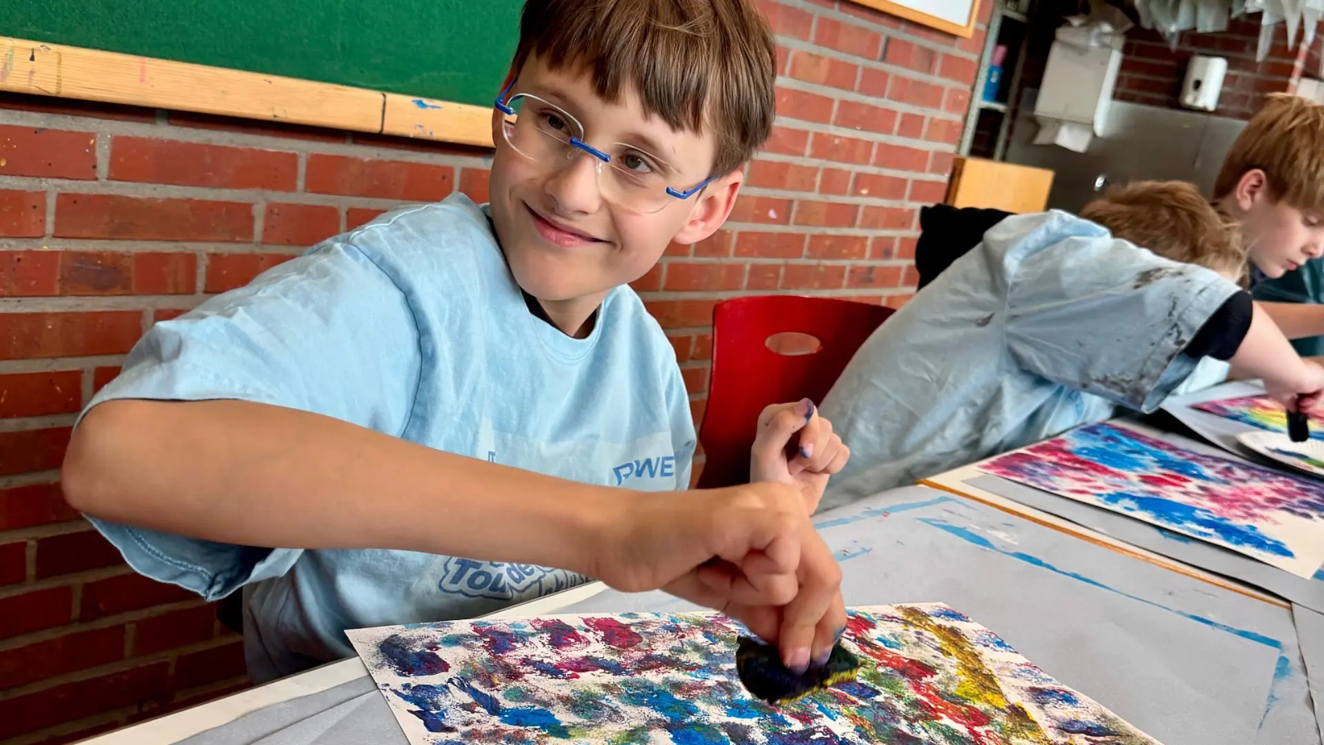 Ein Schüler lächelt fröhlich beim Malen mit Ölfarben. Neben ihm sind auch weitere Schüler*innen zu sehen. 