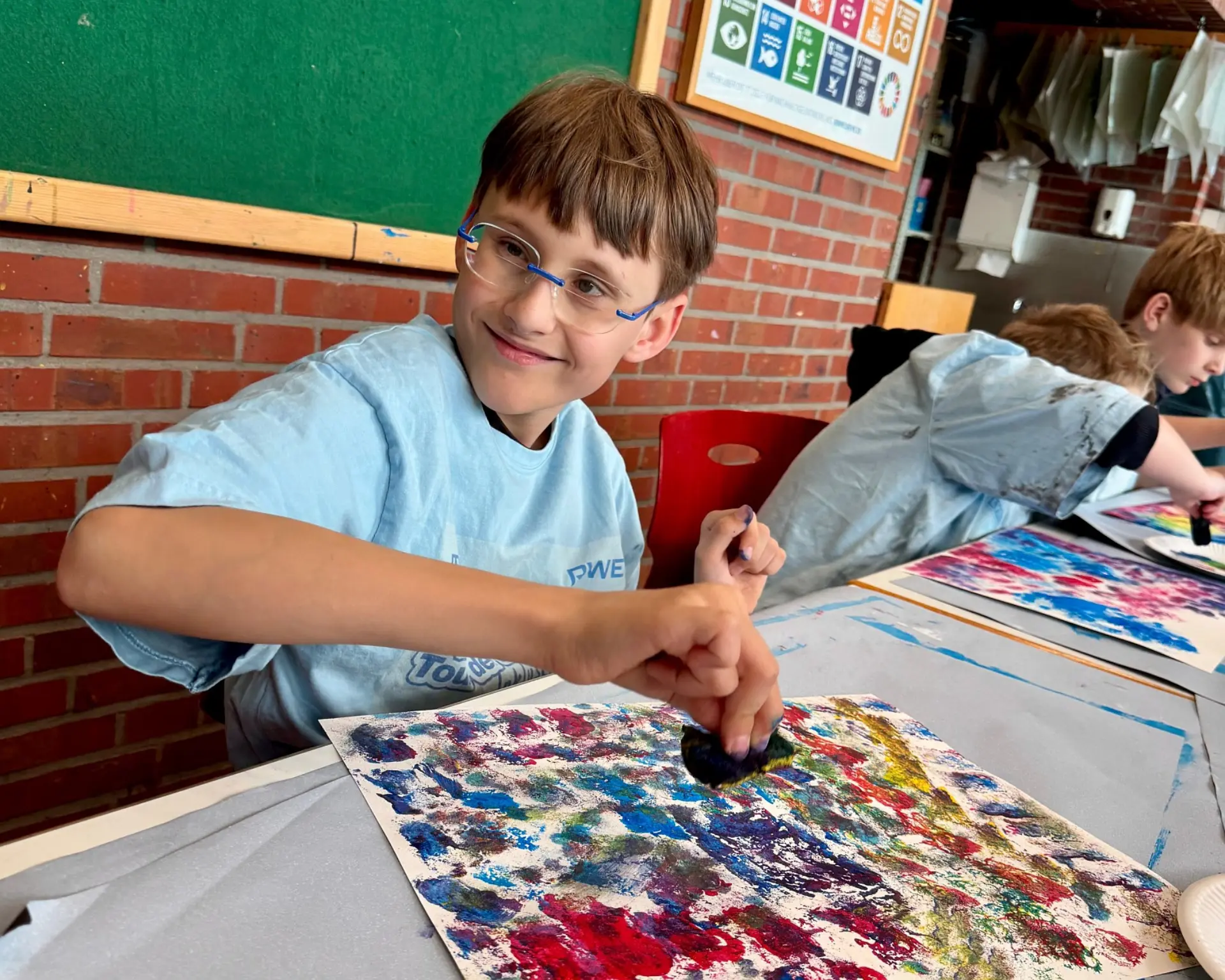 Ein Schüler lächelt fröhlich beim Malen mit Ölfarben. Neben ihm sind auch weitere Schüler*innen zu sehen. 