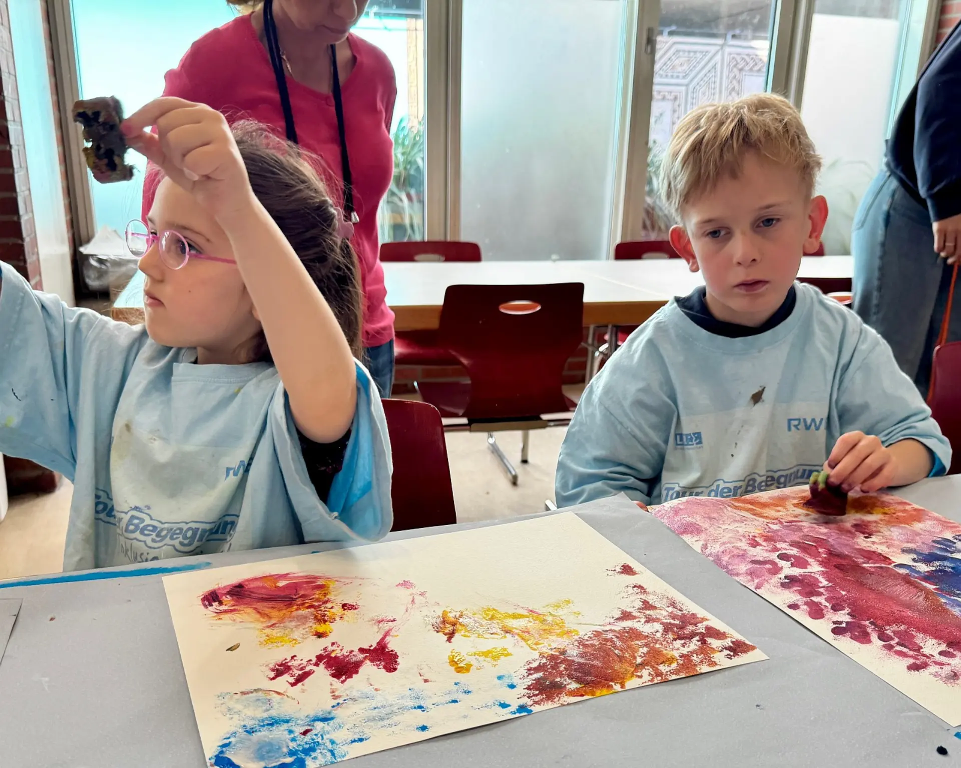 Zwei Schulkinder arbeiten an einem Tisch. Vor ihnen liegen große Blätter, worauf sie mit bunten Aquarellfarben gemalt haben
