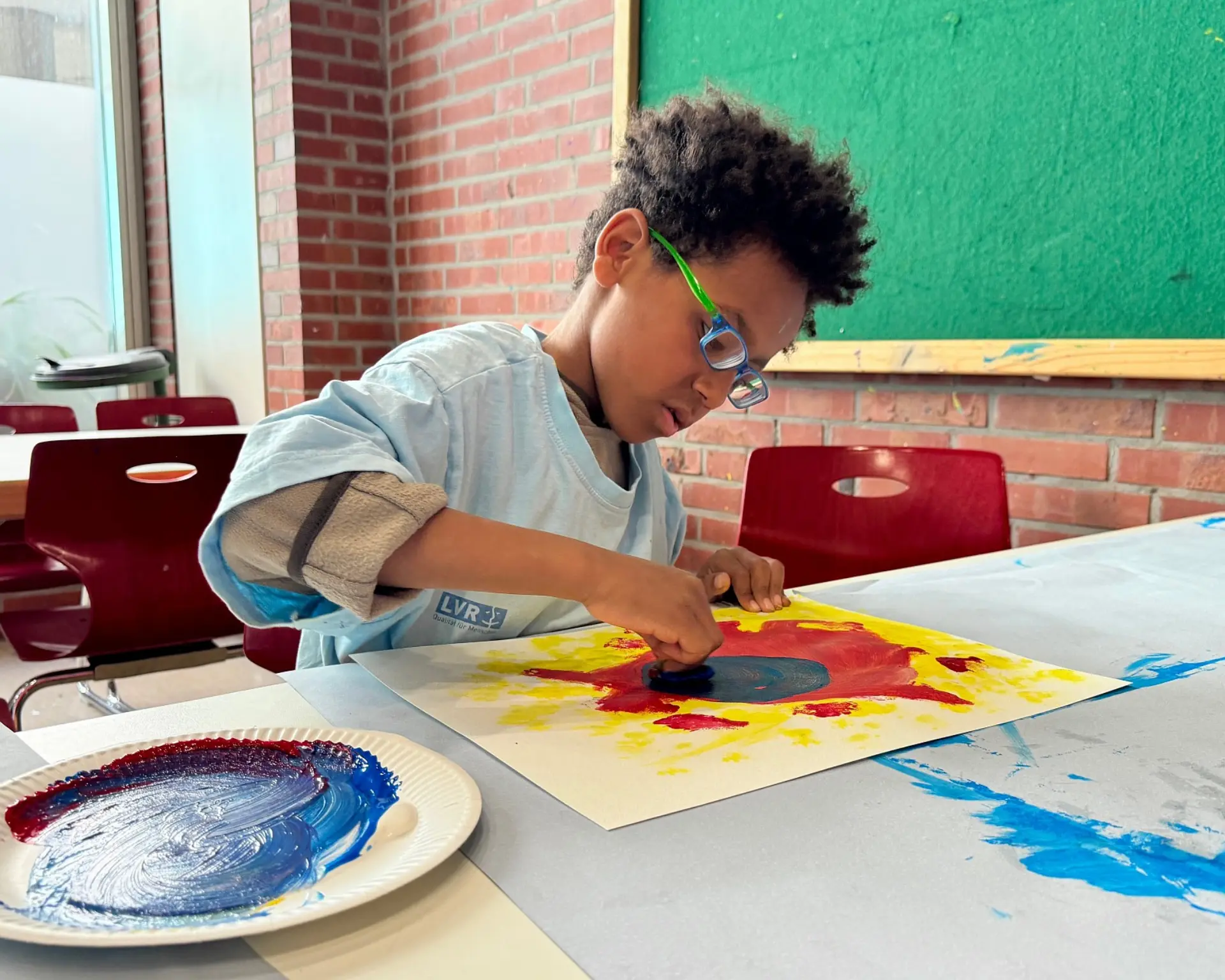 Ein Schulkind sitz am großen Maltisch und mal vertieft mit Aquarellfarben.