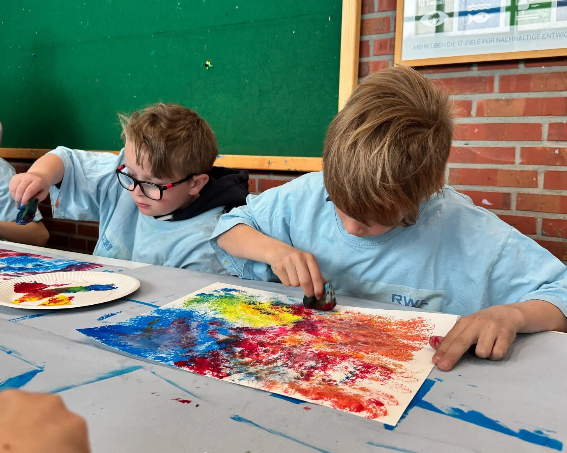 Zwei Schulkinder mit Malschutz-T-Shirts arbeiten fleißig an ihren Aquarellkunstwerken an einem großen Maltisch.