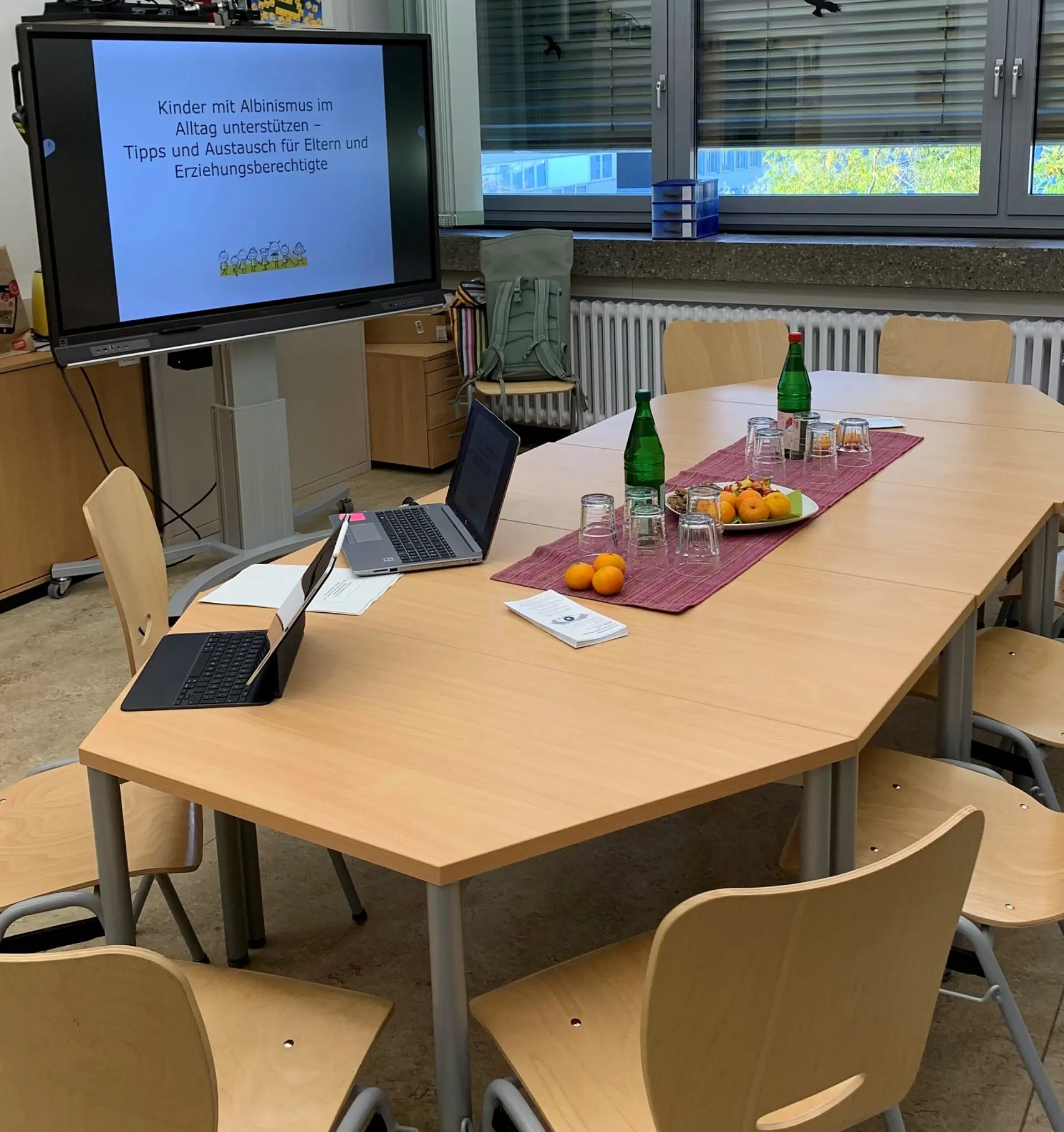 Ein Raum mit einem großen Bildschirm, an dem eine Präsentation gezeigt wird. Davor steht ein langer Tisch mit Stühlen für die Gäste.