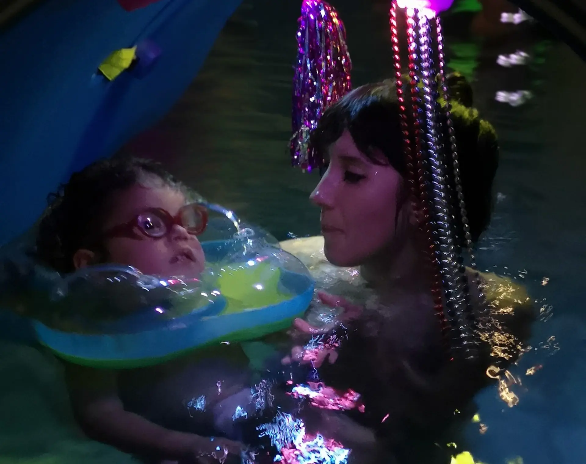 Eine Frau hält ein Kind im Wasser in einem Schwimmbecken. Es liegt im einem Schwimmring. Beide befinden sich in einem bunt ausgeleuchteten Tunnel.