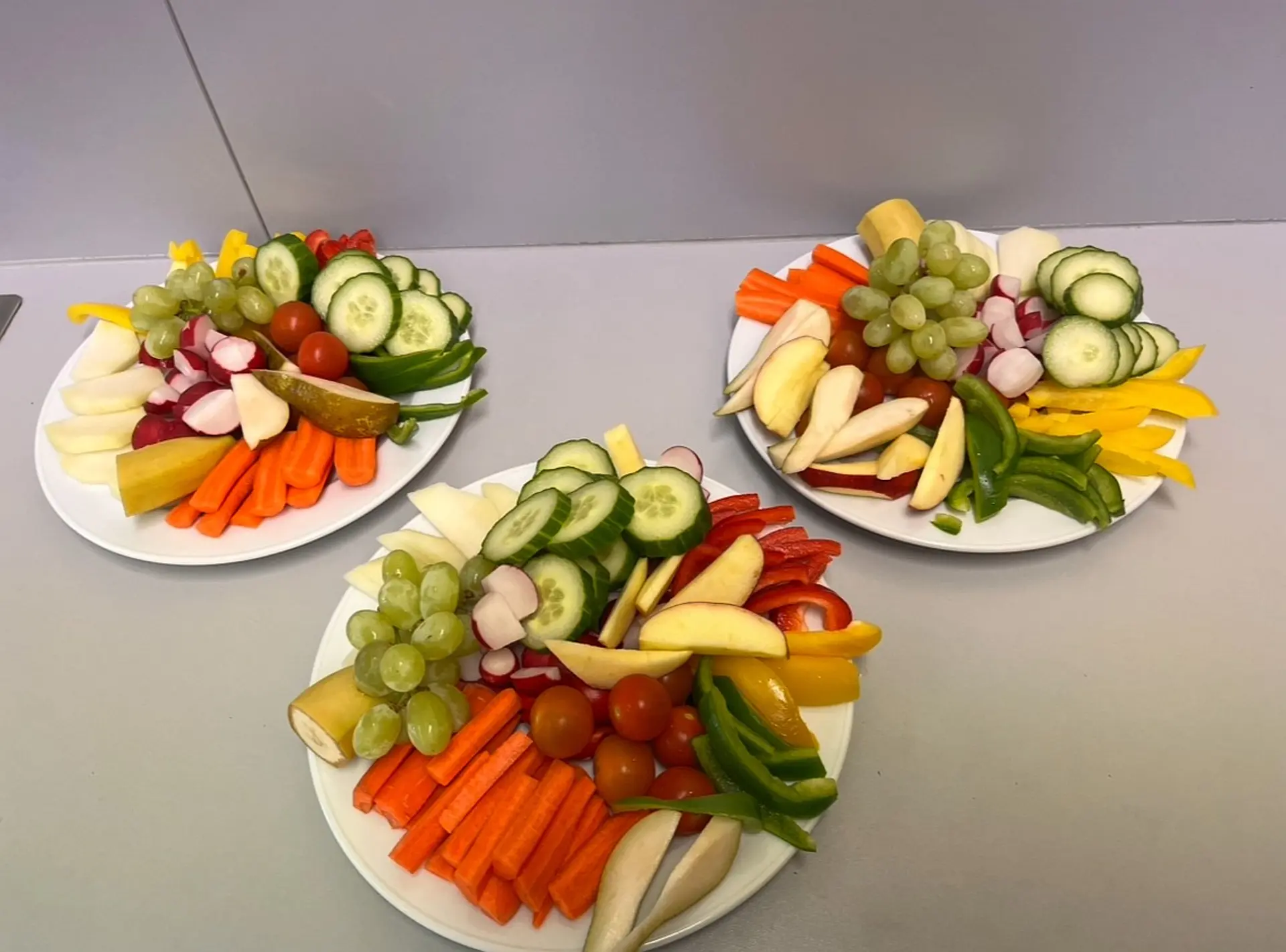 Drei reichhaltig gefüllte weiße Teller mit frischem, buntem Rohkost-Obst und -Gemüse, angeordnet auf der hellgrauen Oberfläche eines Tisches.