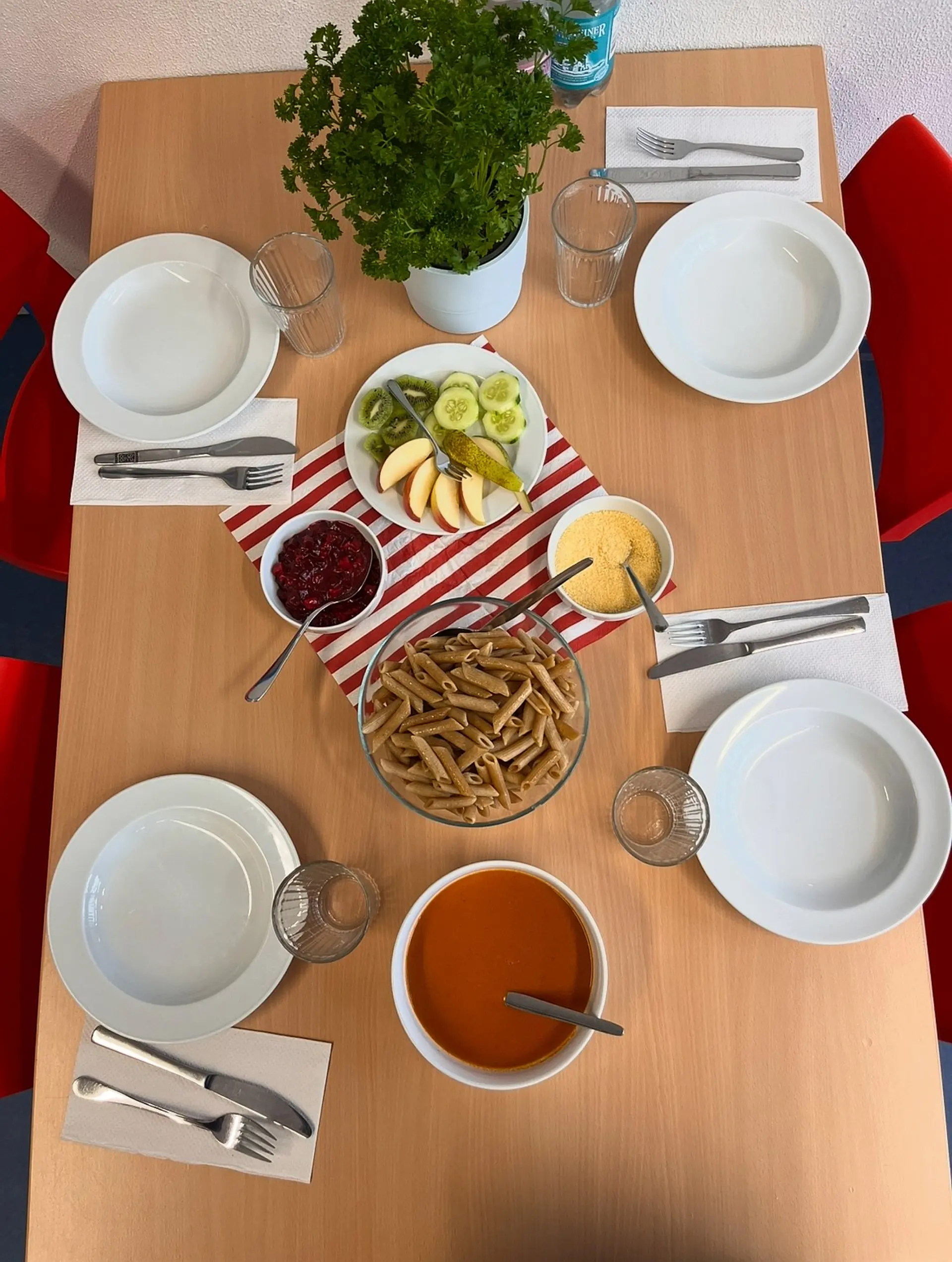 Ein gedeckter Holztisch für vier Personen, vorbereitet für eine Mahlzeit im OGS. Auf dem Tisch stehen vier Teller, Servietten, Besteck und Gläser. In der Mitte sind mehrere Gerichte in Schalen angerichtet: mit Vollkorn-Penne-Nudeln, Tomatensoße, Preiselbeeren-Grütze und geriebenem Käse).