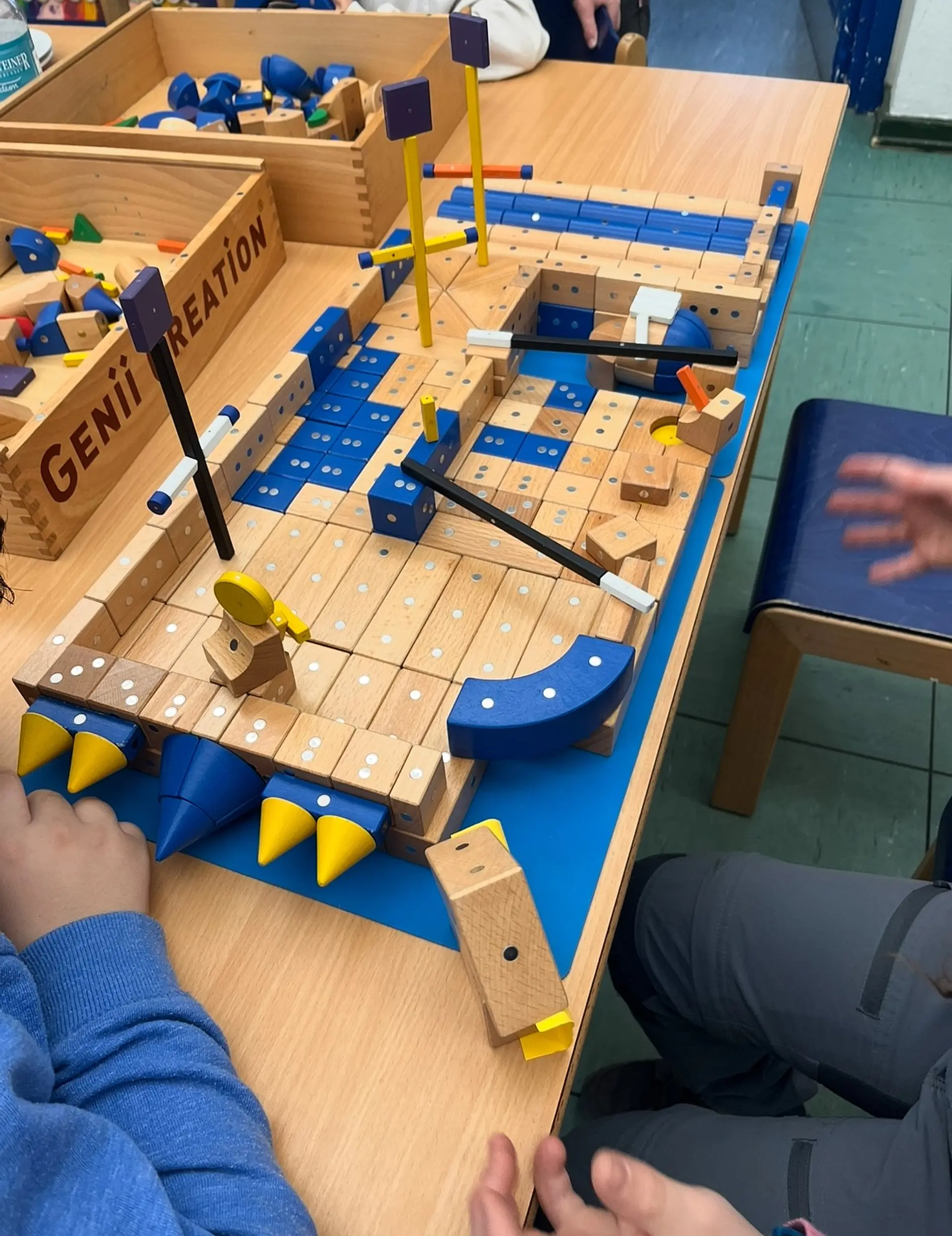Zwei Kinderhände sind an einem Tisch beim Bauen mit einem Satz von hochwertigen, hellen Holzbausteinen (Genii Creation System), die mit Markierungen für blinde und sehbeeinträchtigte Menschen versehen sind..