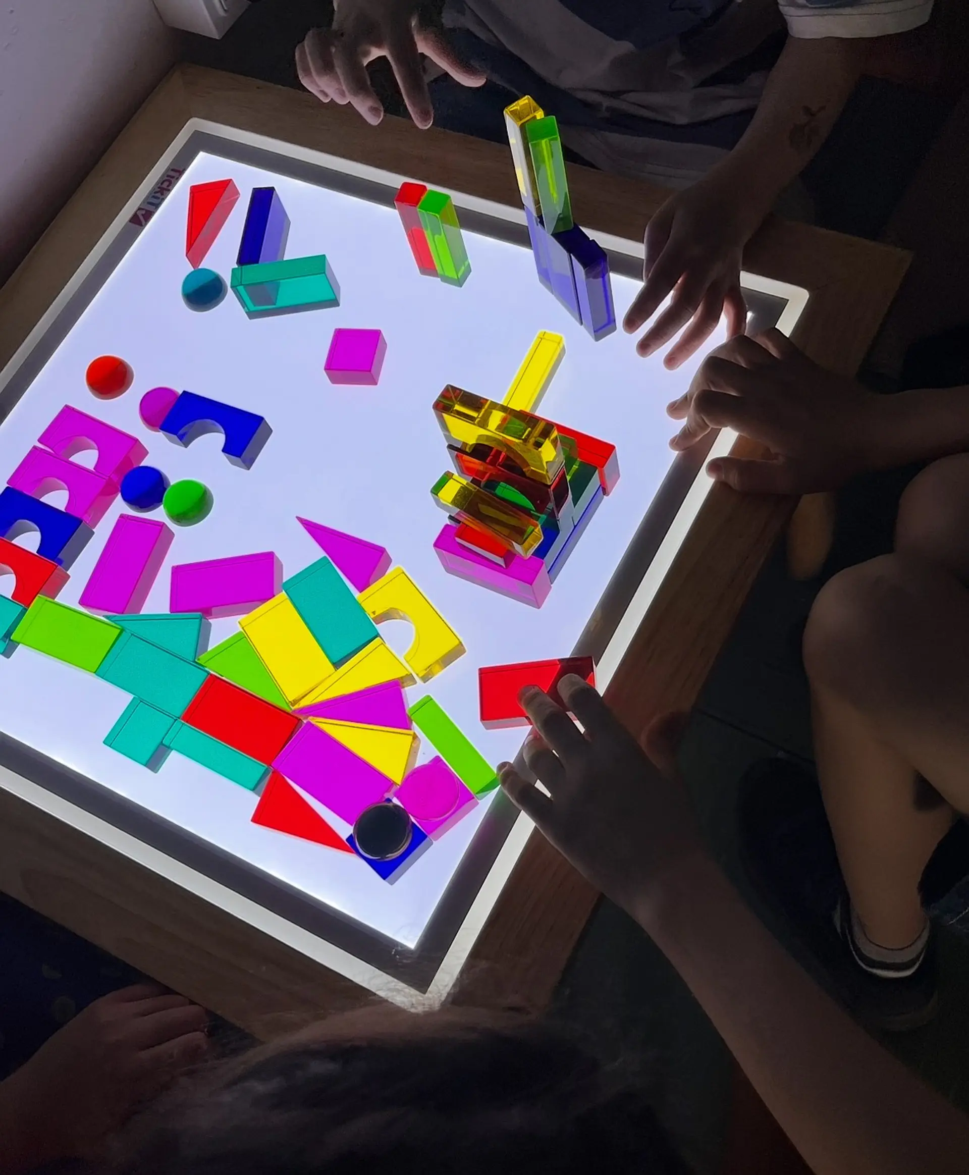 Nahaufnahme eines quadratischen, leuchtenden Lichttisches, auf dem mehrere Kinderhände mit bunten, transparenten Acrylbausteinen spielen. Die Bausteine haben verschiedene geometrische Formen und leuchten in kräftigen Farben.