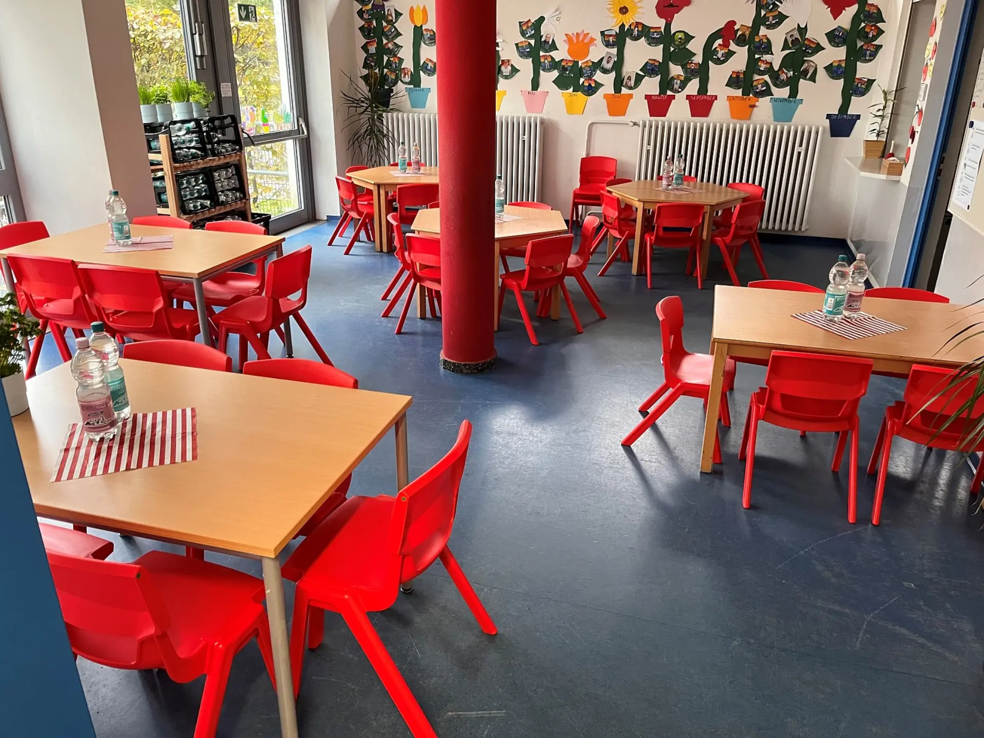 Ein heller, freundlicher Speiseraum, der für das Mittagessen im OGS eingerichtet ist. Im Raum stehen mehrere quadratische Tische mit jeweils vier leuchtend roten Plastikstühlen herum.