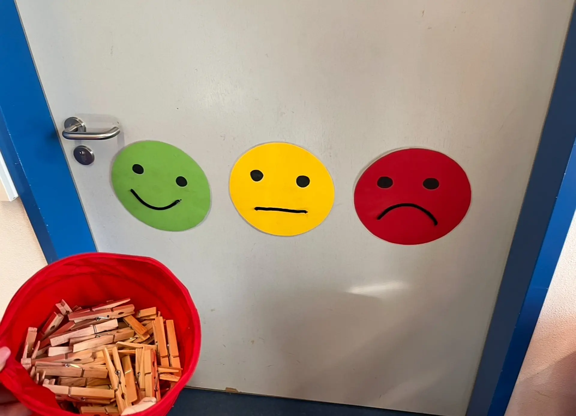 Ein einfaches Stimmungsbarometer an einer Zimmertür. Es besteht aus drei großen, runden Emoticons in Ampelfarben: Links ein grüner Smiley (fröhlich), in der Mitte ein gelber Smiley (neutral/nachdenklich) und rechts ein roter Smiley (traurig/verärgert). Unten hält eine Hand einen roten Eimer voller hölzerner Wäscheklammern bereit, die die Kinder nutzen, um ihre aktuelle Stimmung auf der Skala anzuzeigen.