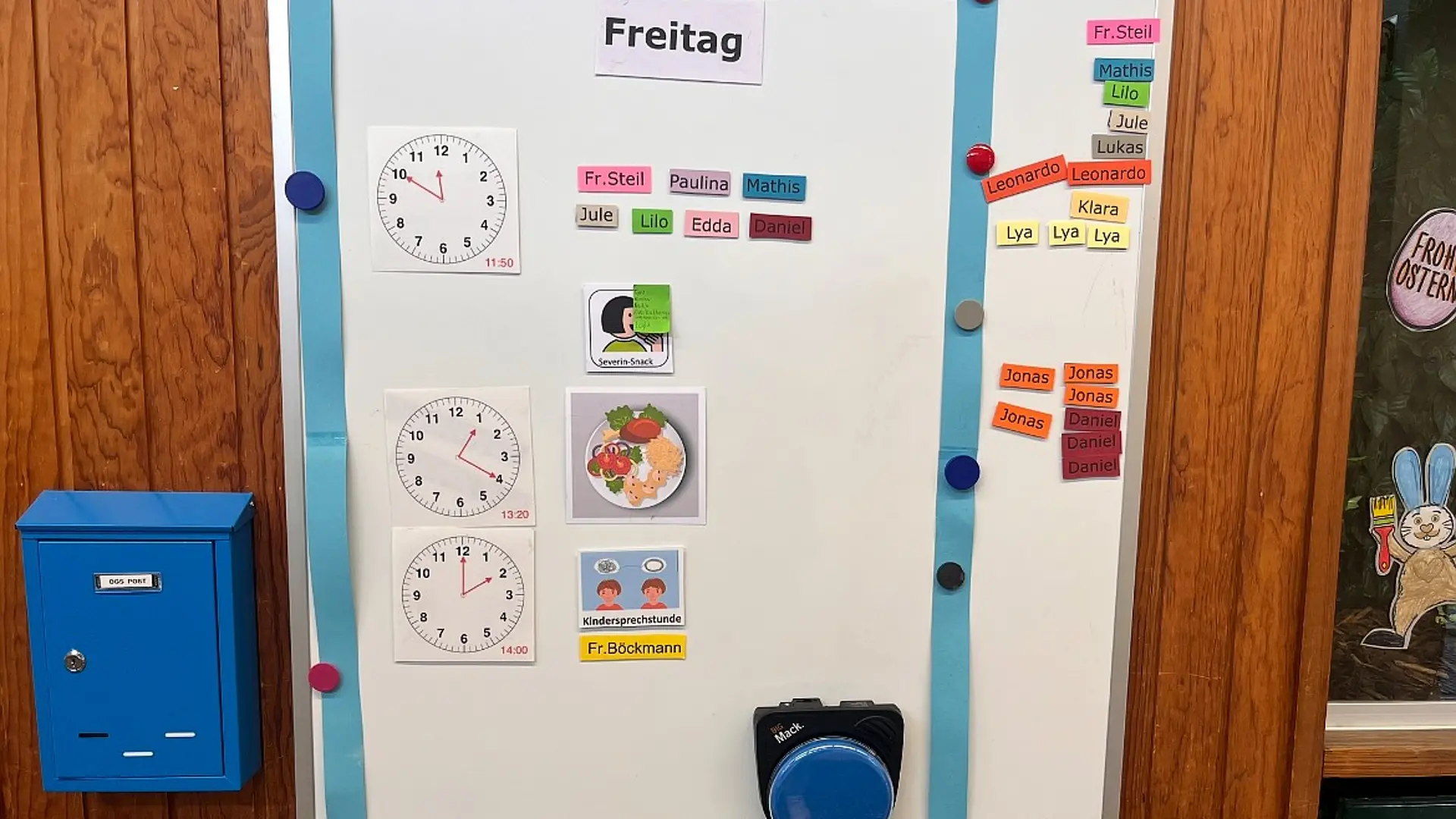 Ein Tagesplan für einen Freitag, montiert an einer Holzwand, der die Struktur des Offenen Ganztags visualisiert. Der Plan verwendet analoge Uhren, Bildkarten und Namensschilder. Links hängt ein kleiner Briefkasten mit der Aufschrift 'POST'.