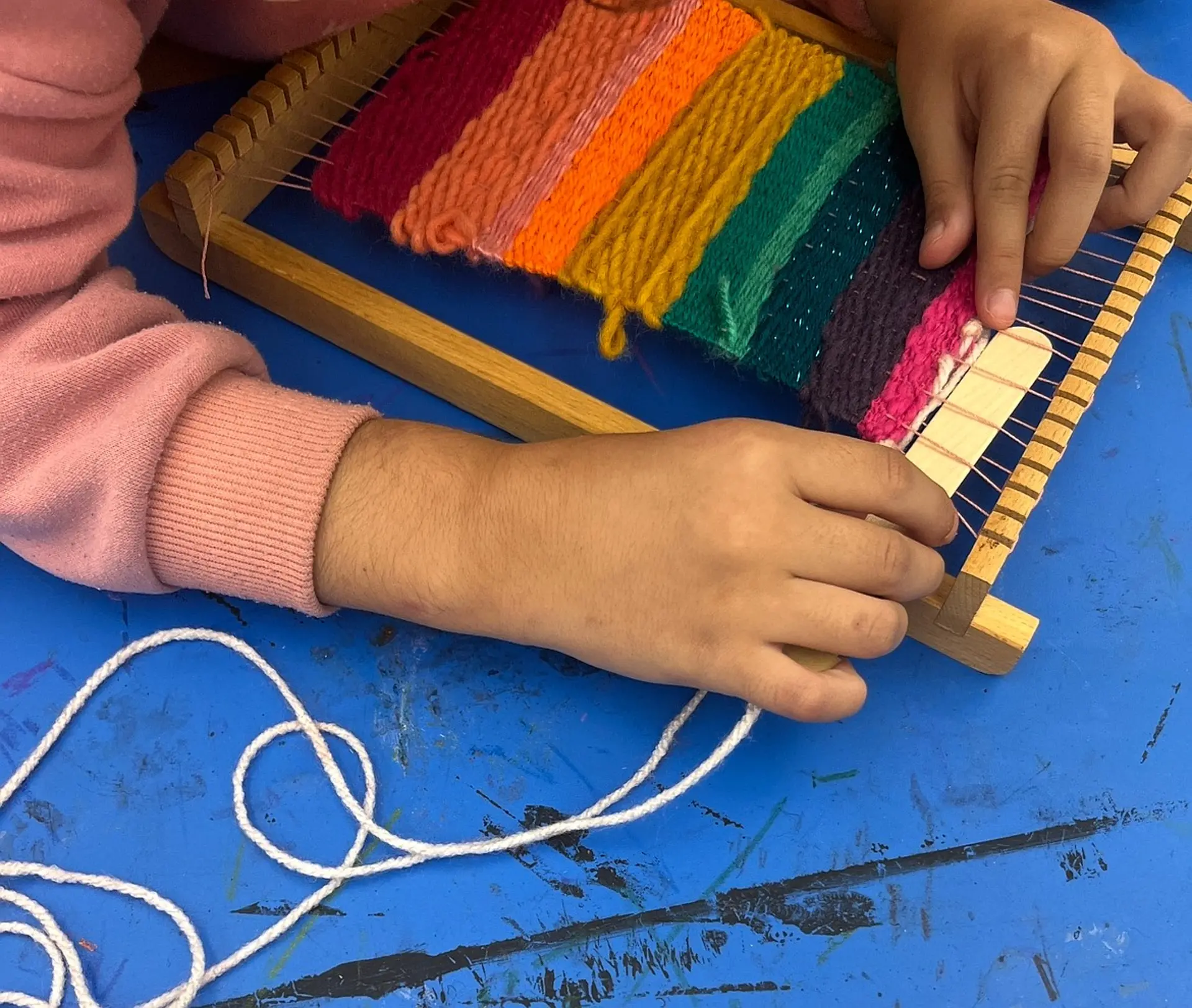 Nahaufnahme von zwei Kinderhänden, die an einem kleinen, rechteckigen Holzwebrahmen arbeiten. Das Kind webt farbenfrohe Wollfäden in leuchtenden Regenbogenfarben ein.
