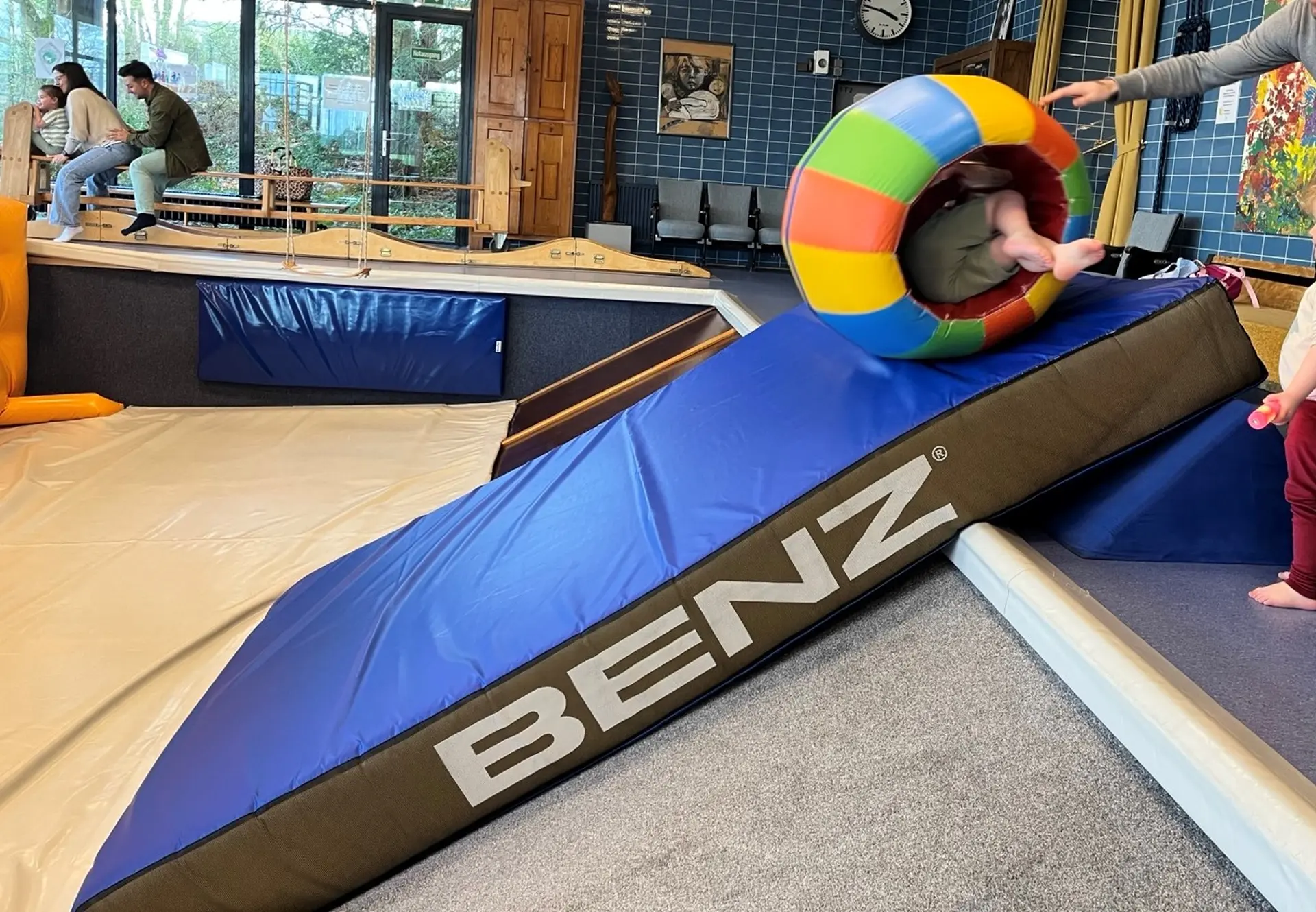 Das Bild zeigt eine bunte Rolle mit einem Kind, dass eine Matte hinab rollt. Im Hintergrund ist eine Familie auf einer Wippe.