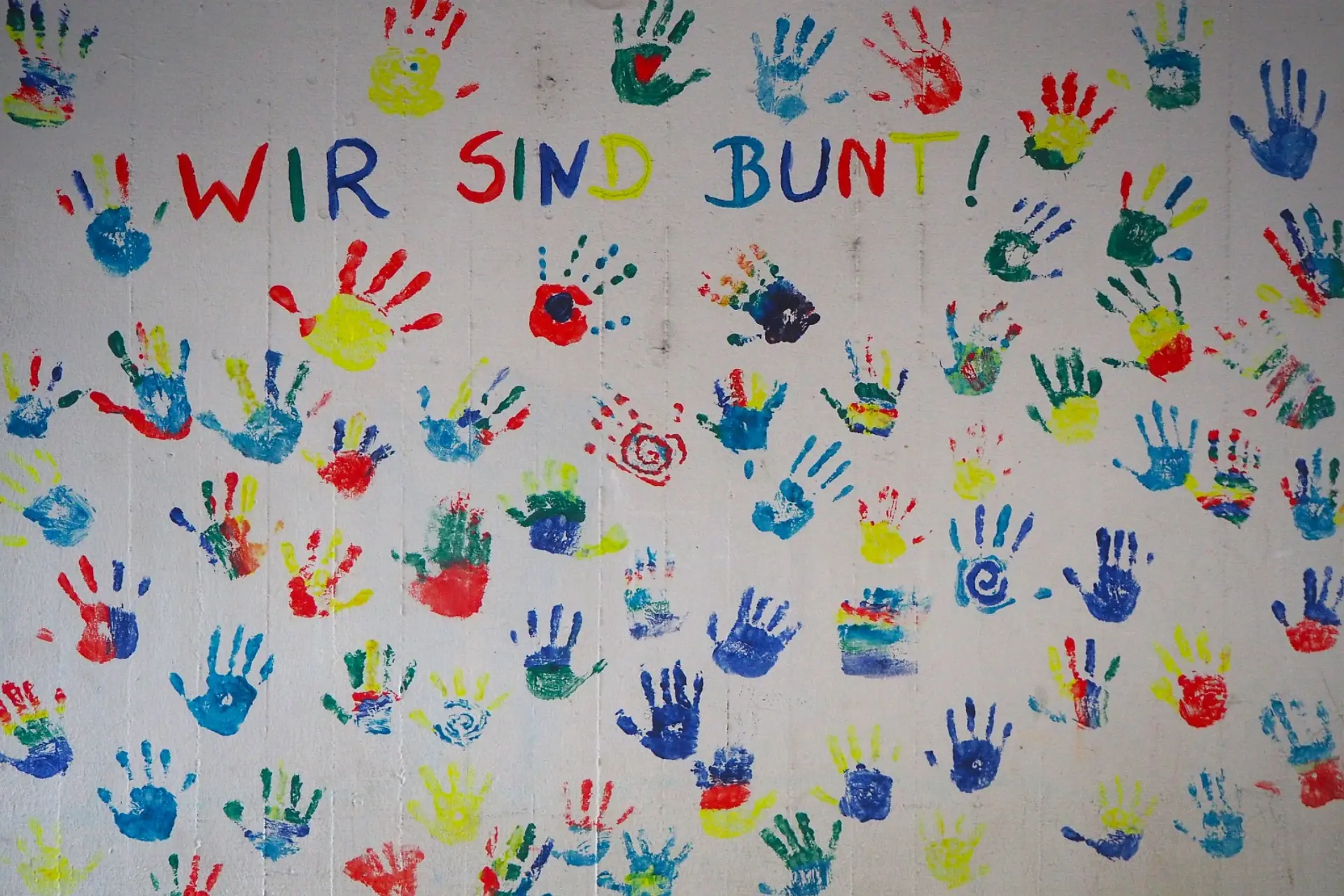 Eine Schulwand mit Duzenden von Kinderhandabdrücken in bunten Farben. An der Wand ist auch Wir sind bunt geschrieben.