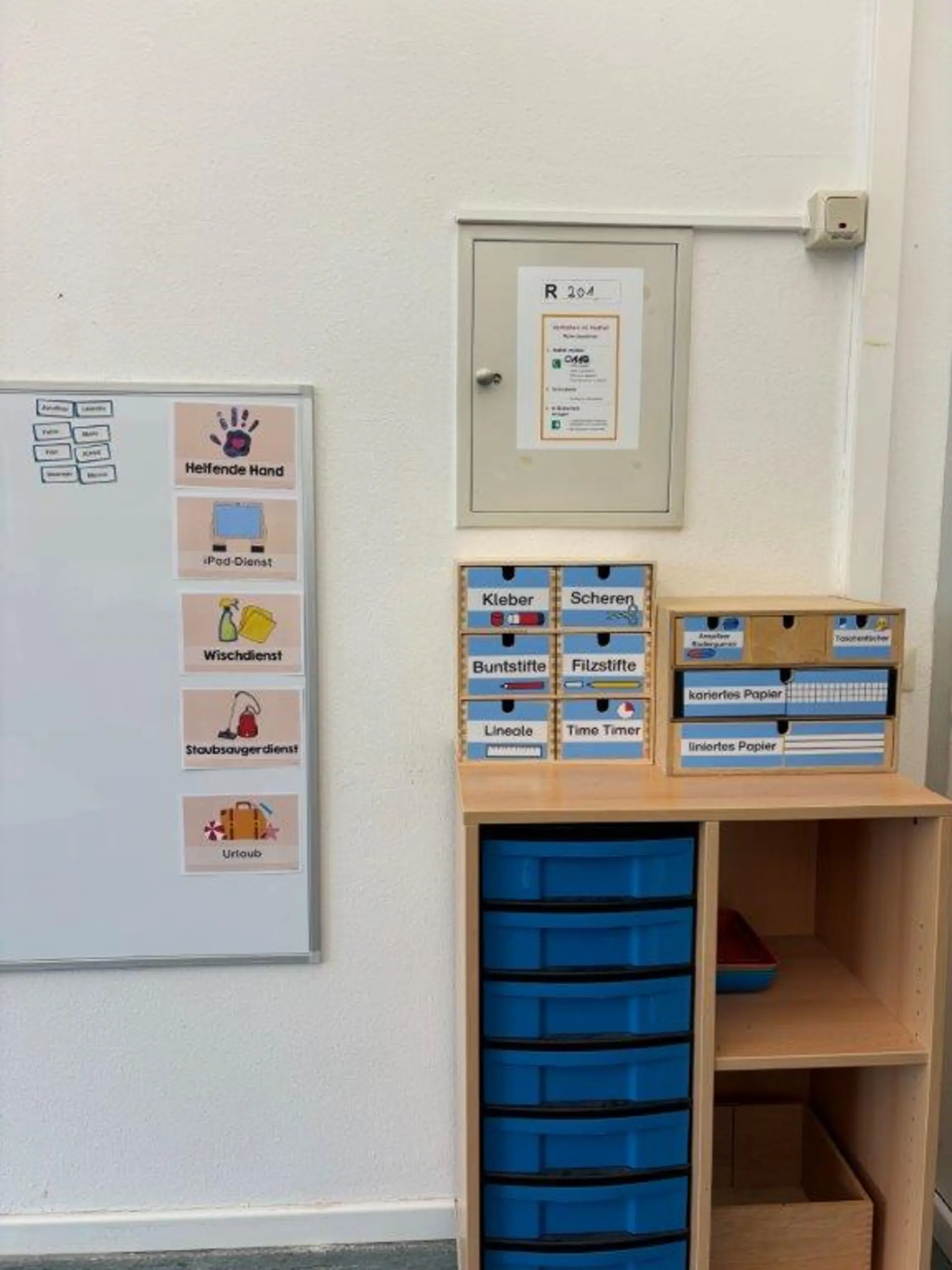 Ein aufgeräumter Bereich im Klassenraum mit einer Schreibtafel, auf der illustrierte Kärtchen die Klassendienste (Helferhand, iPad-Dienst, Wischdienst, Staubsaugerdienst, Urlaub) visualisieren. Rechts davon stehen Holzregale mit Schubladen, die beschriftete Materialien wie Kleber, Scheren, Buntstifte und liniertes bzw. kariertes Papier enthalten.