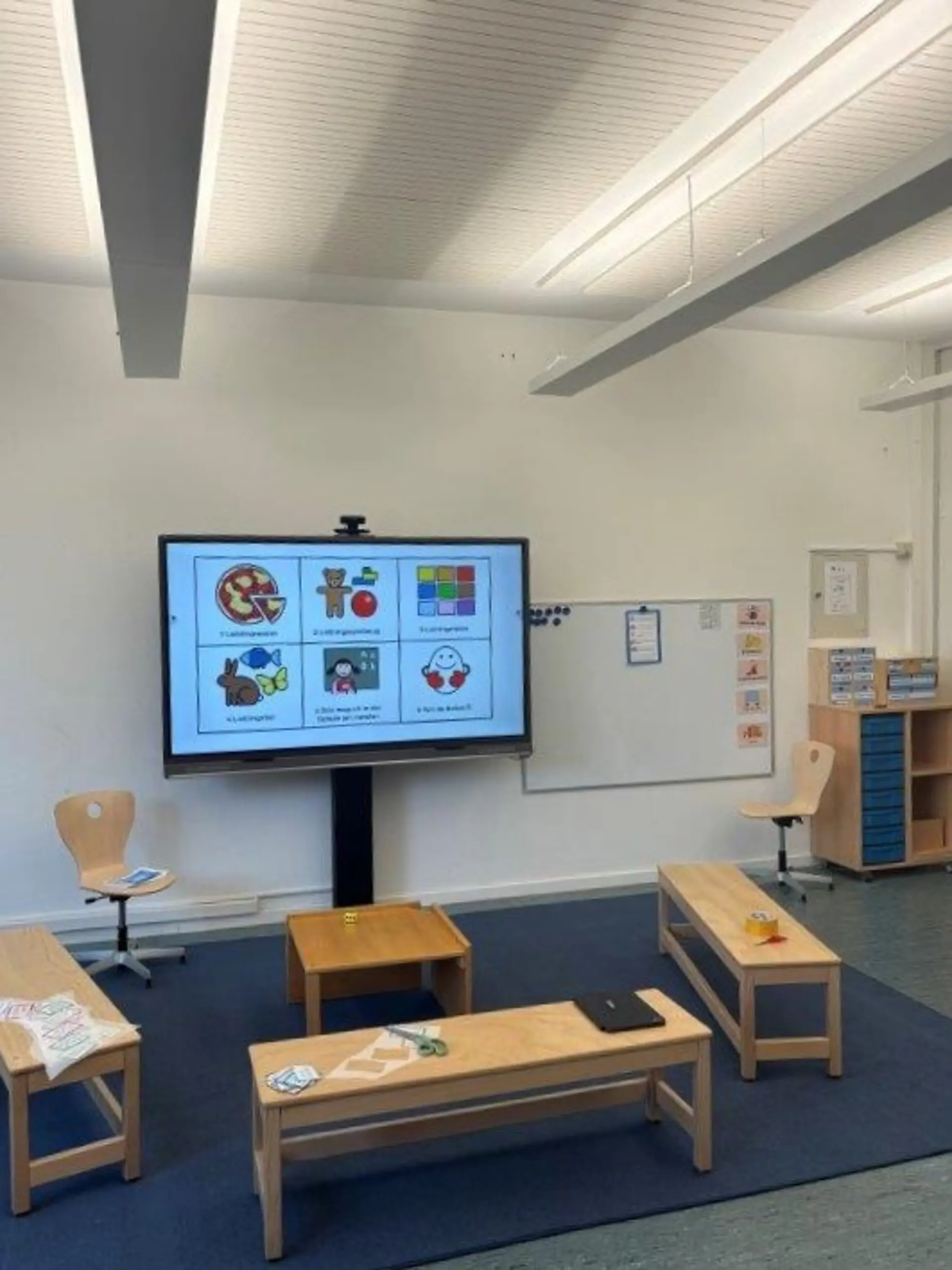Ein modern ausgestatteter Klassenraum: In der Mitte steht ein großer interaktiver Monitor, der sechs bildliche Symbole mit Aufgaben oder Regeln anzeigt. Davor und daneben sind niedrige, flexible Holzbänke und ein kleiner Tisch in U-Form auf einem Teppich platziert, um einen Sitzkreis zu bilden. Im Hintergrund sind ein Whiteboard und niedrige Regale zu sehen.