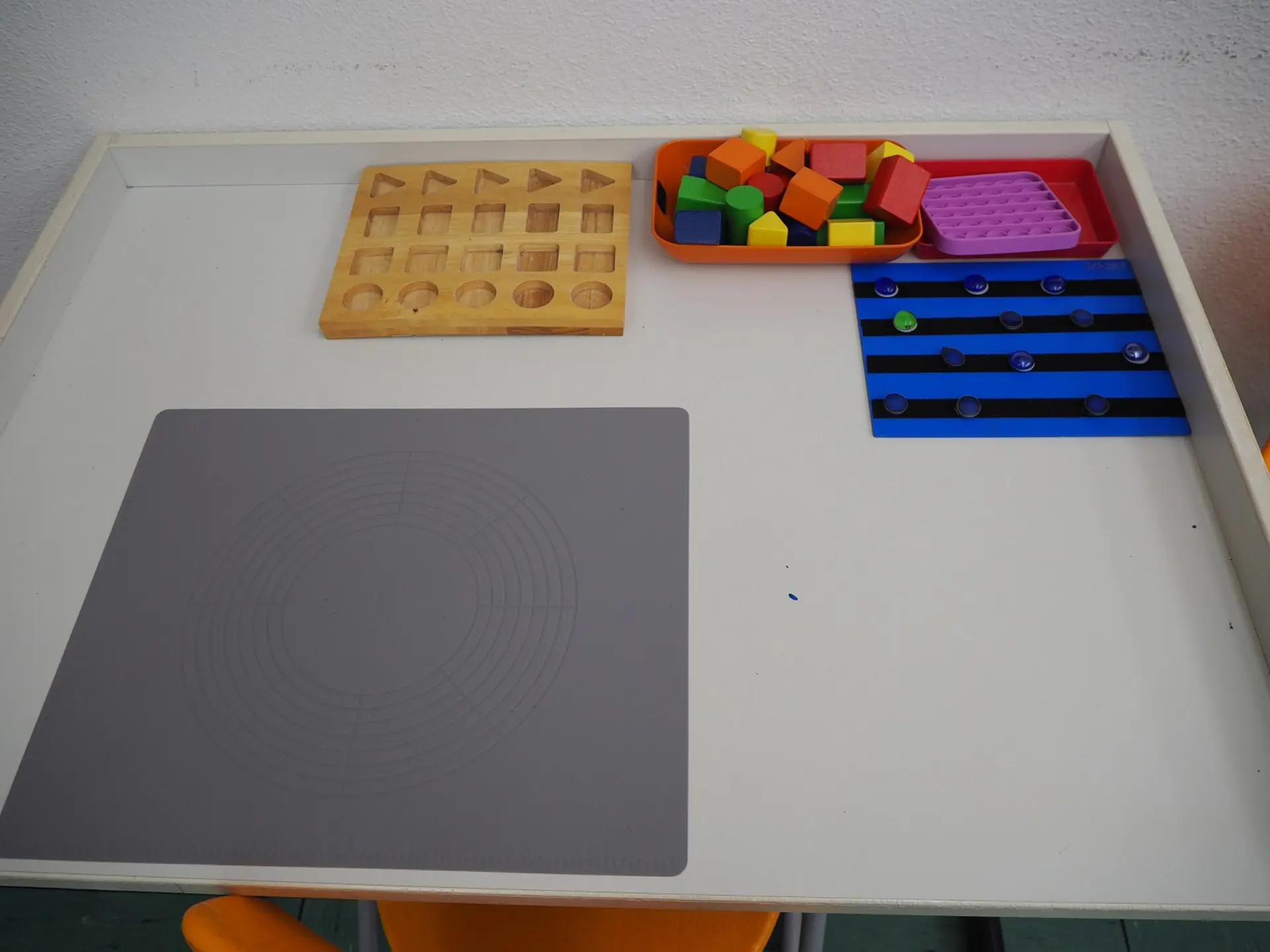 Arbeitsplatz für taktiles und visuelles Lernen: Auf einem Tisch liegen verschiedene Fördermaterialien: Eine Holztafel mit ausgeschnittenen geometrischen Formen, ein Korb mit bunten Bauklötzen und einer Pop-It-Platte sowie eine Platte mit taktilen Markierungen und Noppen. Links liegt eine große Matte mit kreisförmigen, zentrischen Linien.