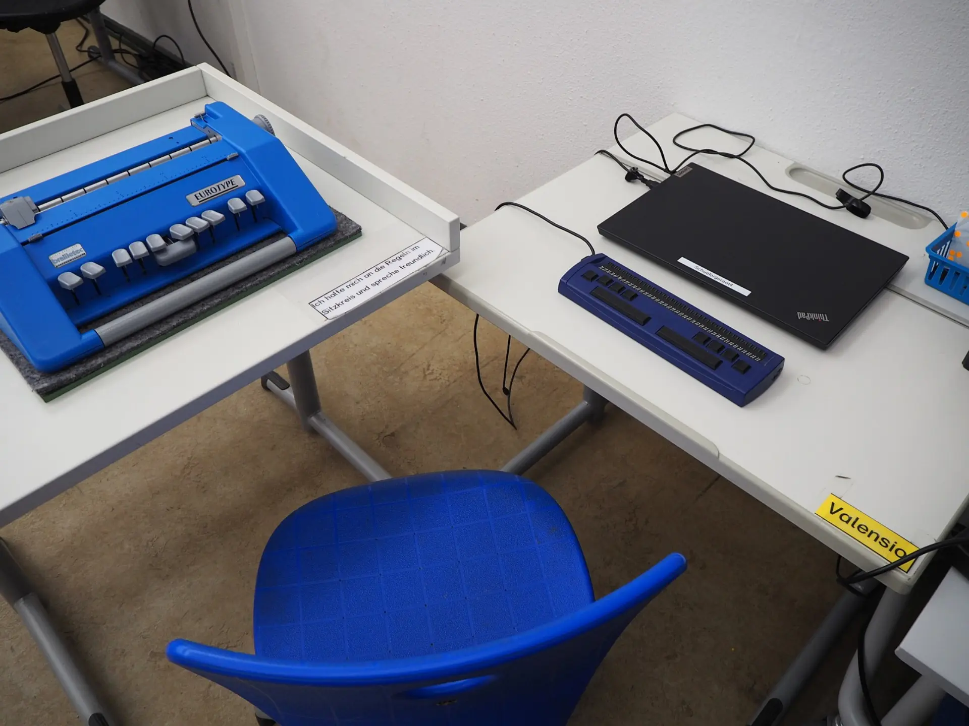 Arbeitsplatz für blinde und sehbeeinträchtigte Schüler*innen: Links steht eine mechanische Braille-Schreibmaschine. Rechts liegt eine Braille-Tastatur, die mit einem Laptop verbunden ist. Auf dem Tisch liegt ein Zettel mit der Aufschrift: Ich helfe euch mit den Fingern zu lesen und zu schreiben.