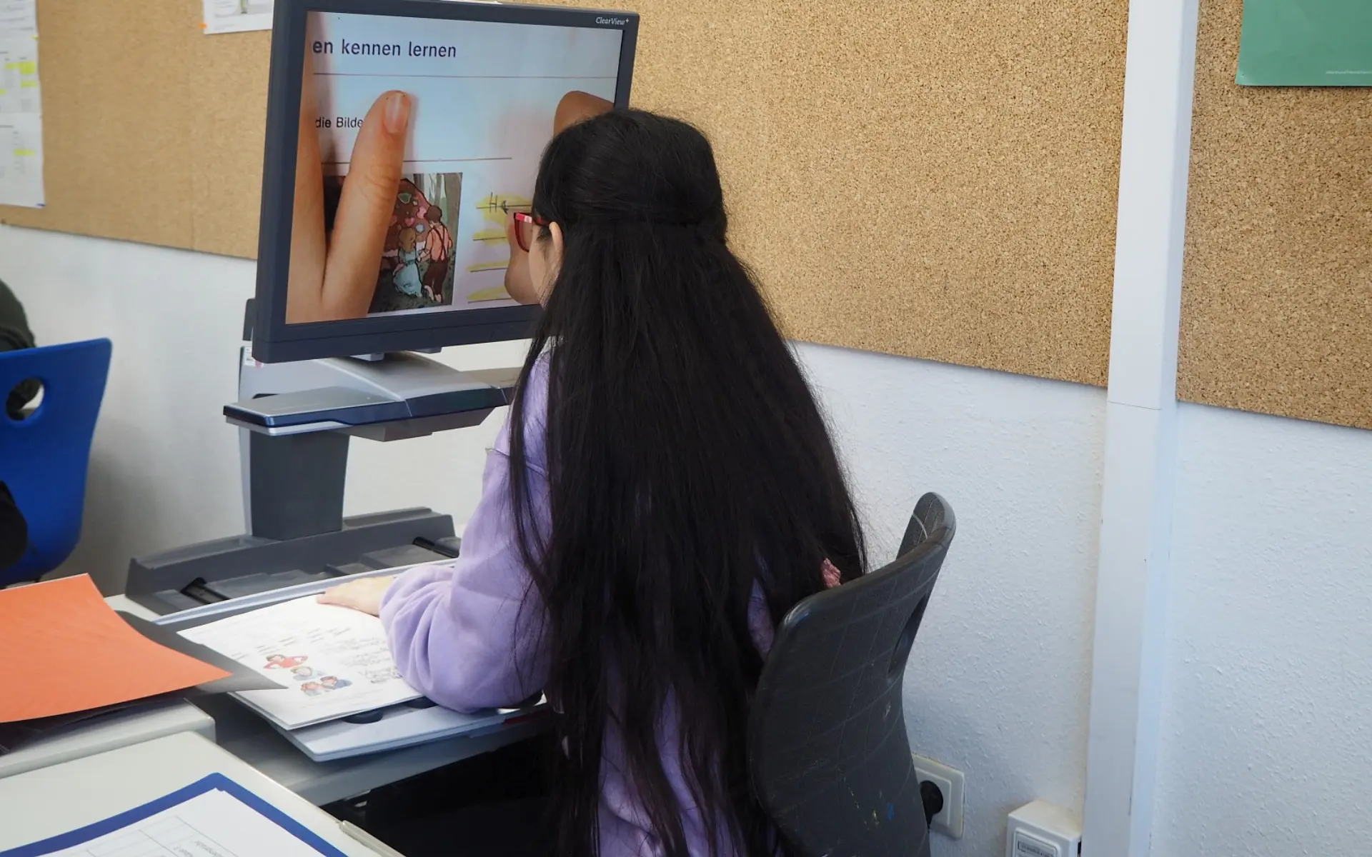 Eine Schülerin arbeitet an einem Schreibtisch, der mit einem Bildschirmlesegerät ausgestattet ist. Sie schaut ganz nah auf den Monitor, wo sie groß angezeigt bekommt, wie sie Buchstaben schreiben übt.