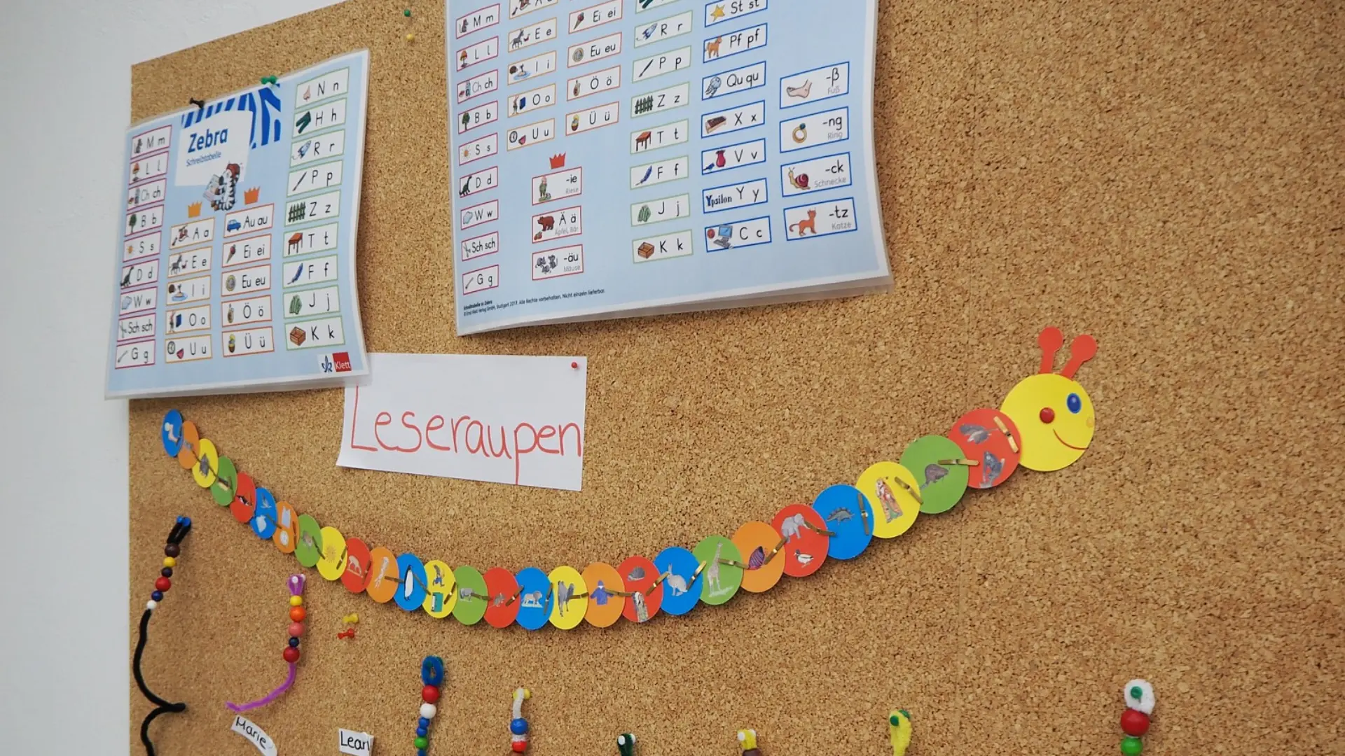 Pinnwand in der Primarstufe: Oben hängen zwei Plakate mit Buchstaben und Anlauten zur Leseförderung. Darunter ist eine lange, bunte Leseraupe aus Pappkreisen angebracht. Mehrere kurze, geperlte Schnüre hängen von der Raupe herunter, die mit den Namen einzelner Schüler*innen beschriftet sind.
