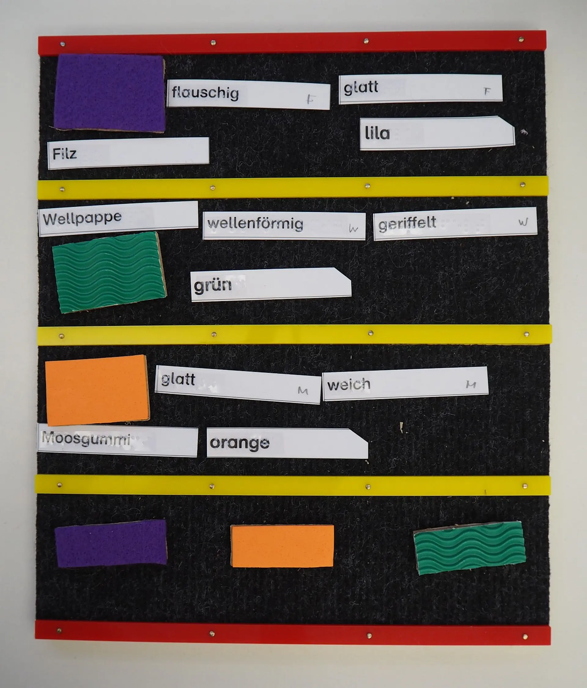 Eine Filztafel, die taktiles Zuordnungsmaterial zeigt. Verschiedene Stoff- und Moosgummi-Rechtecke müssen den entsprechenden Schildern wie Filz, Moosgummi, Wellpappe, flauschig, glatt, lila und orange zugeordnet werden.