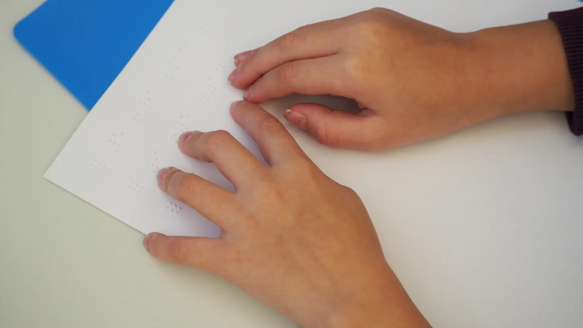 Nahaufnahme von zwei Kinderhänden, die sanft über ein Blatt Papier tasten, auf dem Blindenschrift in erhabenen Punkten geprägt ist. Das Bild zeigt das taktile Lesen von Braille.